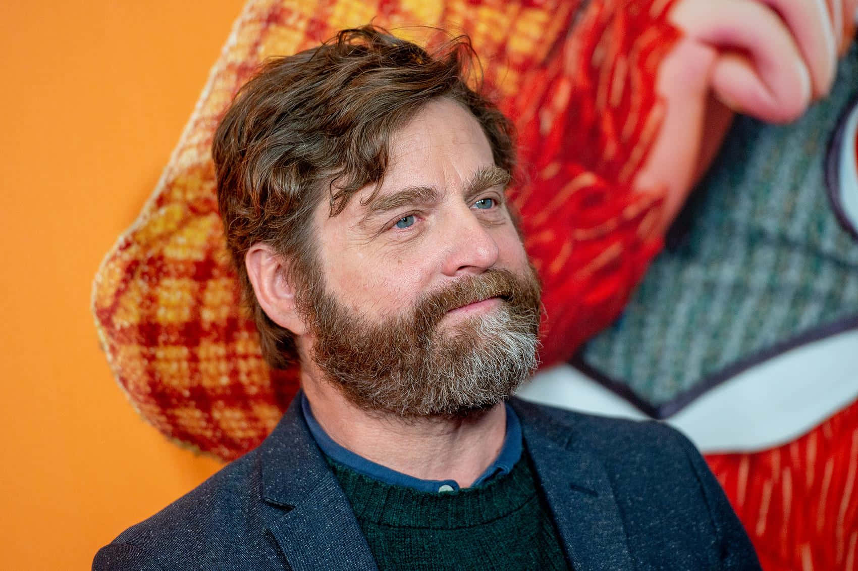 Zach Galifianakis 桌布 壁紙 — 一個留著濃密鬍鬚、藍眼睛的男人，在充滿活力的橙色和紅色圖案背景下若有所思地凝視著。
