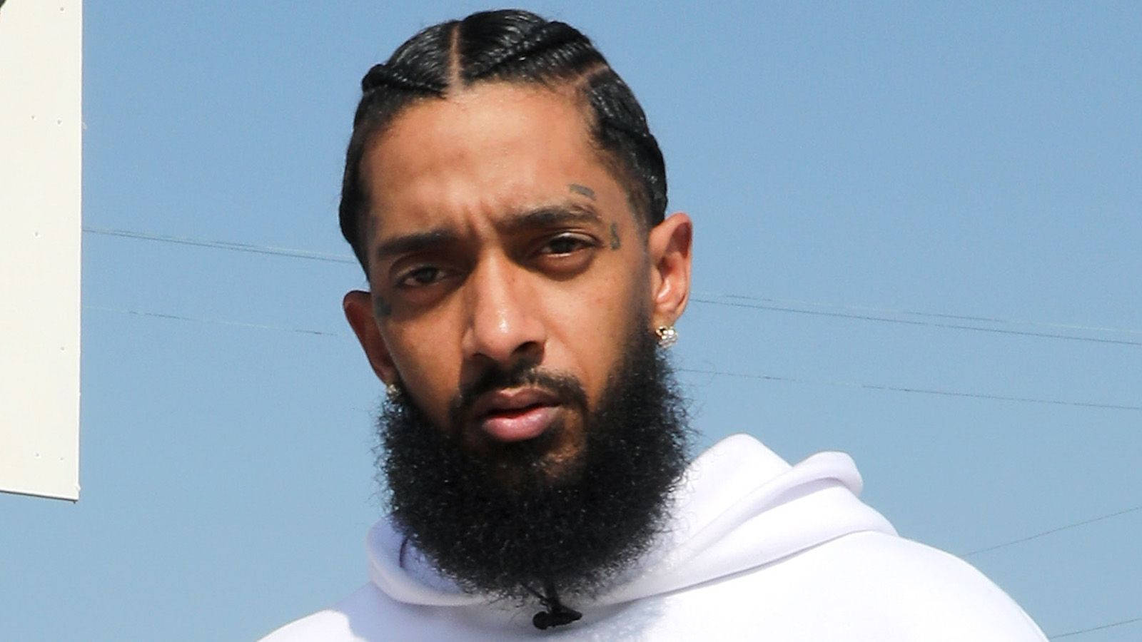 Nipsey Hussle 桌布 壁紙 — 一個留著濃密鬍鬚和玉米辮的男人，穿著白色連帽衫，在清澈的藍天下凝視著。