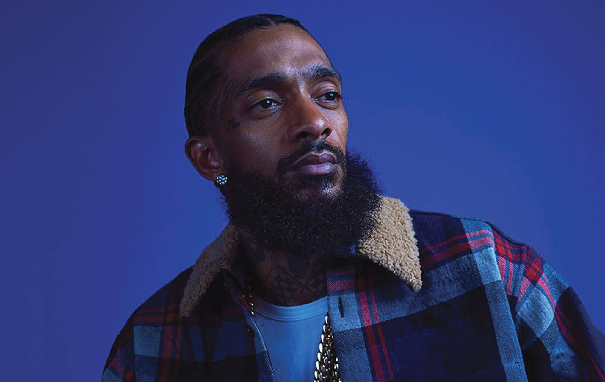 Nipsey Hussle 桌布 壁紙 — 一個留著濃密鬍鬚和紋身的男人，穿著格子外套，背景是純藍色。