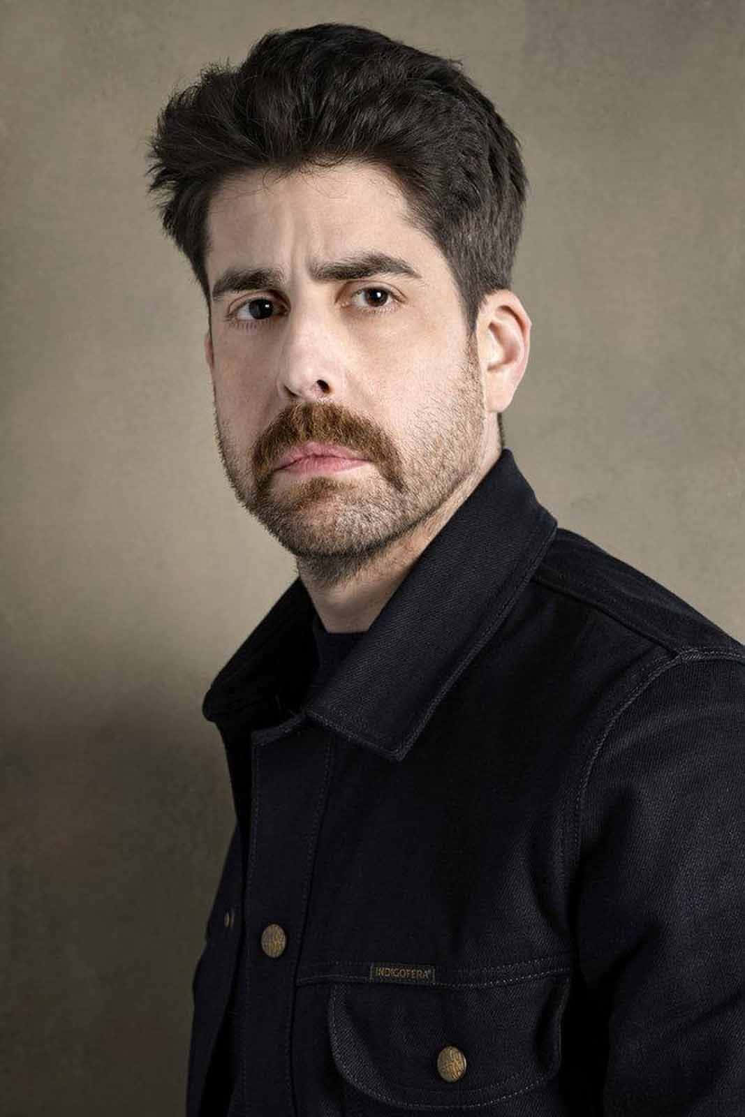 actor 桌布 壁紙 — 一個留著濃密鬍鬚和鬍子的男人，穿著深色牛仔外套，在柔和的灰色背景下專注地凝視鏡頭。