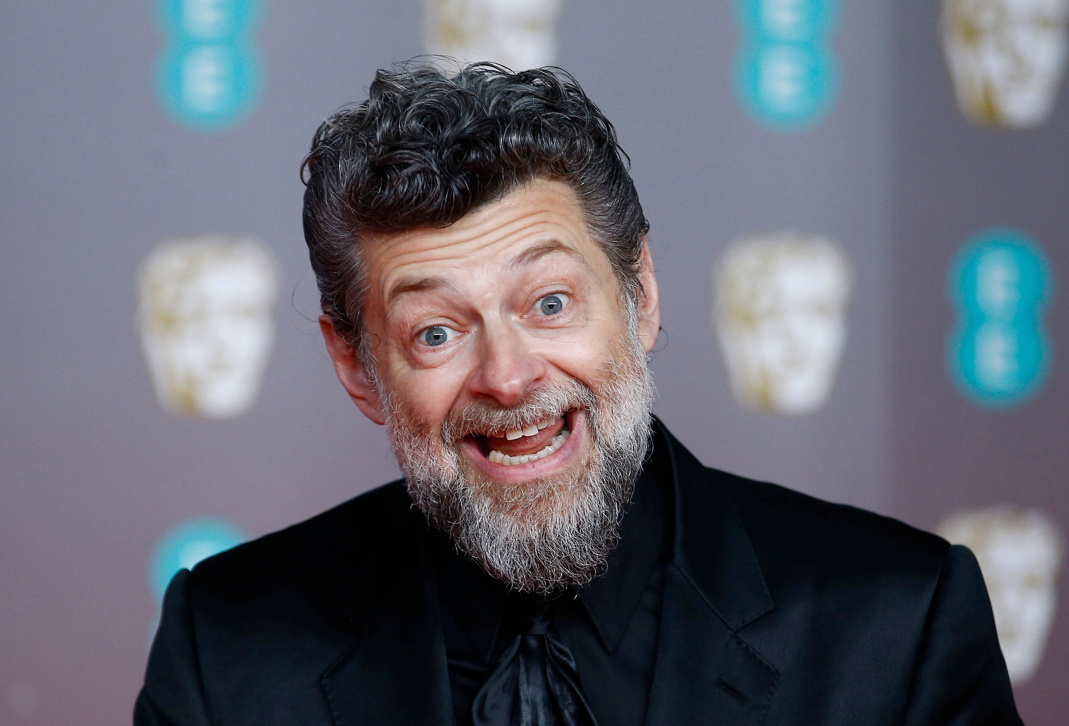 actor 桌布 壁紙 — 一個留著灰白鬍子、頭髮蓬亂的男人，在重複的BAFTA面具背景下對著鏡頭咧嘴大笑。