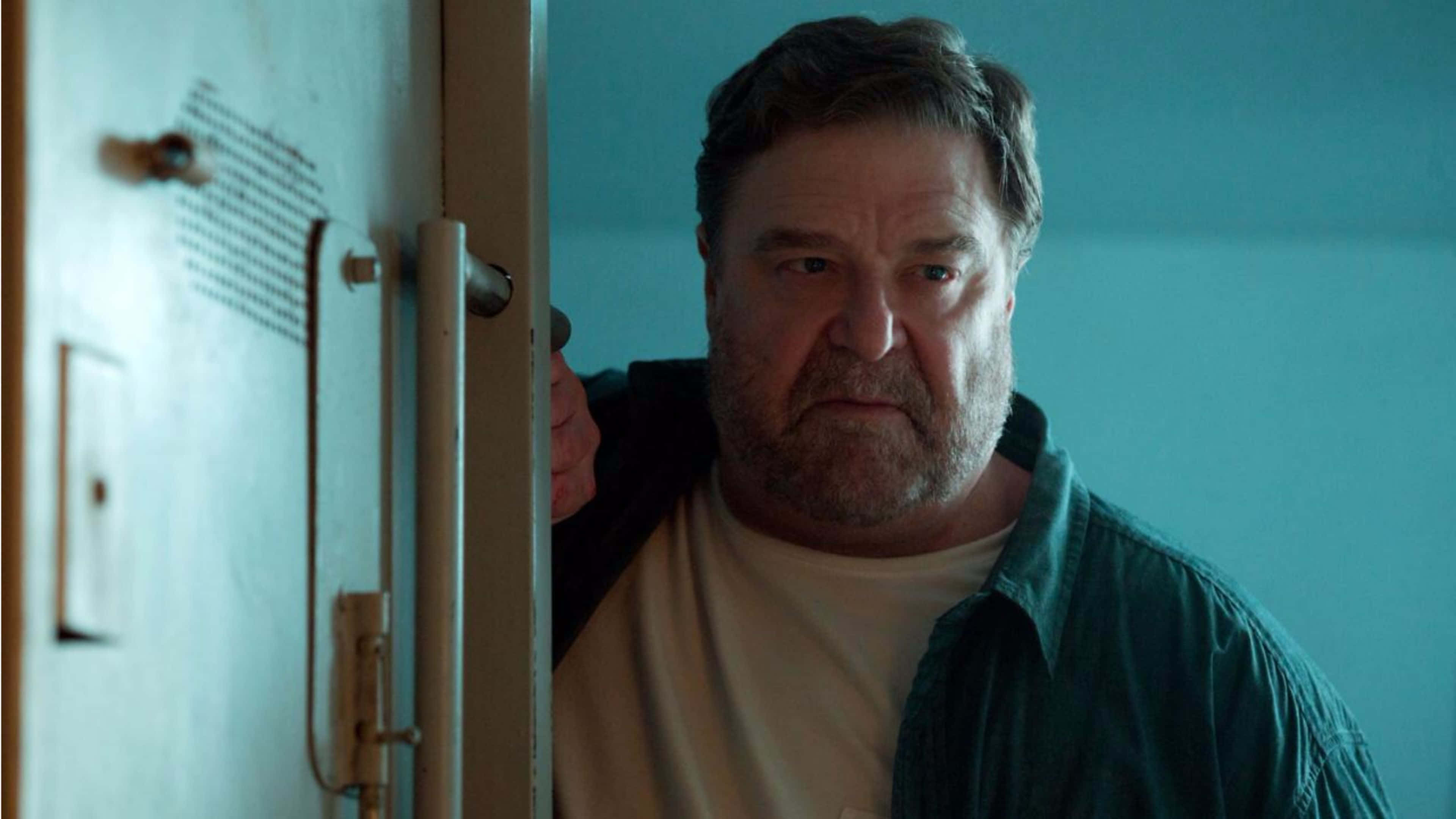 John Goodman 桌布 壁紙 — 一個留著灰白鬍鬚的男人，在燈光昏暗、牆壁呈青綠色的房間裡，小心翼翼地從金屬門後窺視。