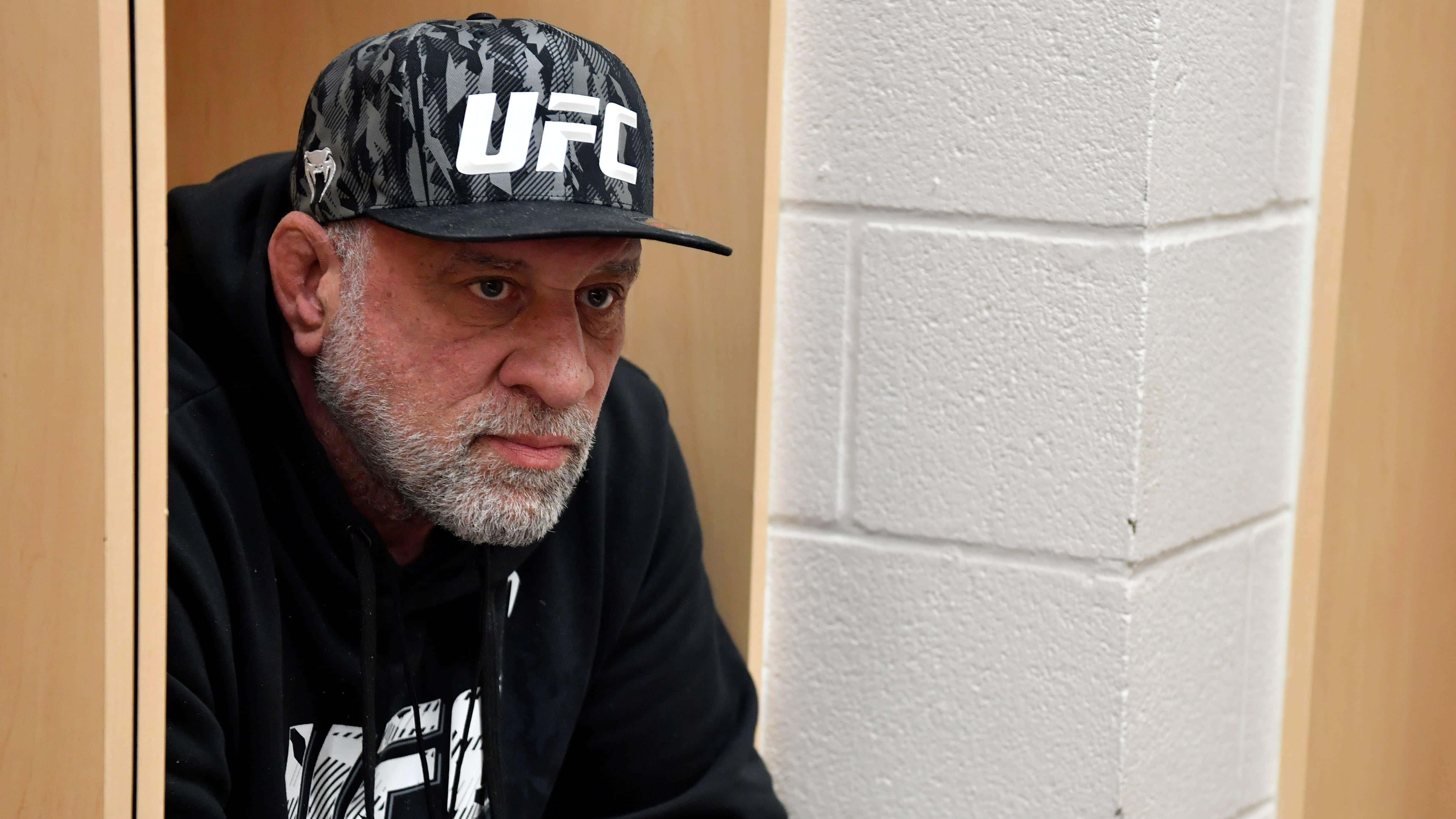 UFC 桌布 壁紙 — 一個留著灰色鬍鬚、戴著UFC帽子的嚴肅男人，從木門之間窺視，背景是白色磚牆。