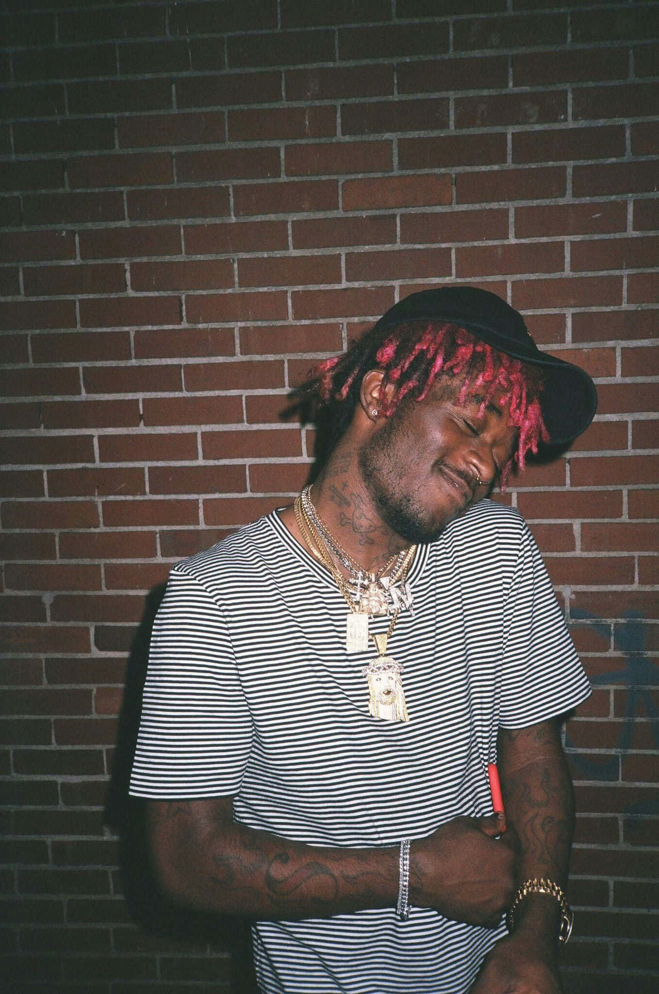 21 Savage 桌布 壁紙 — 一個留著粉紅色辮子、戴著金鏈的男人在晚上靠著磚牆，閉著眼睛，姿勢放鬆。