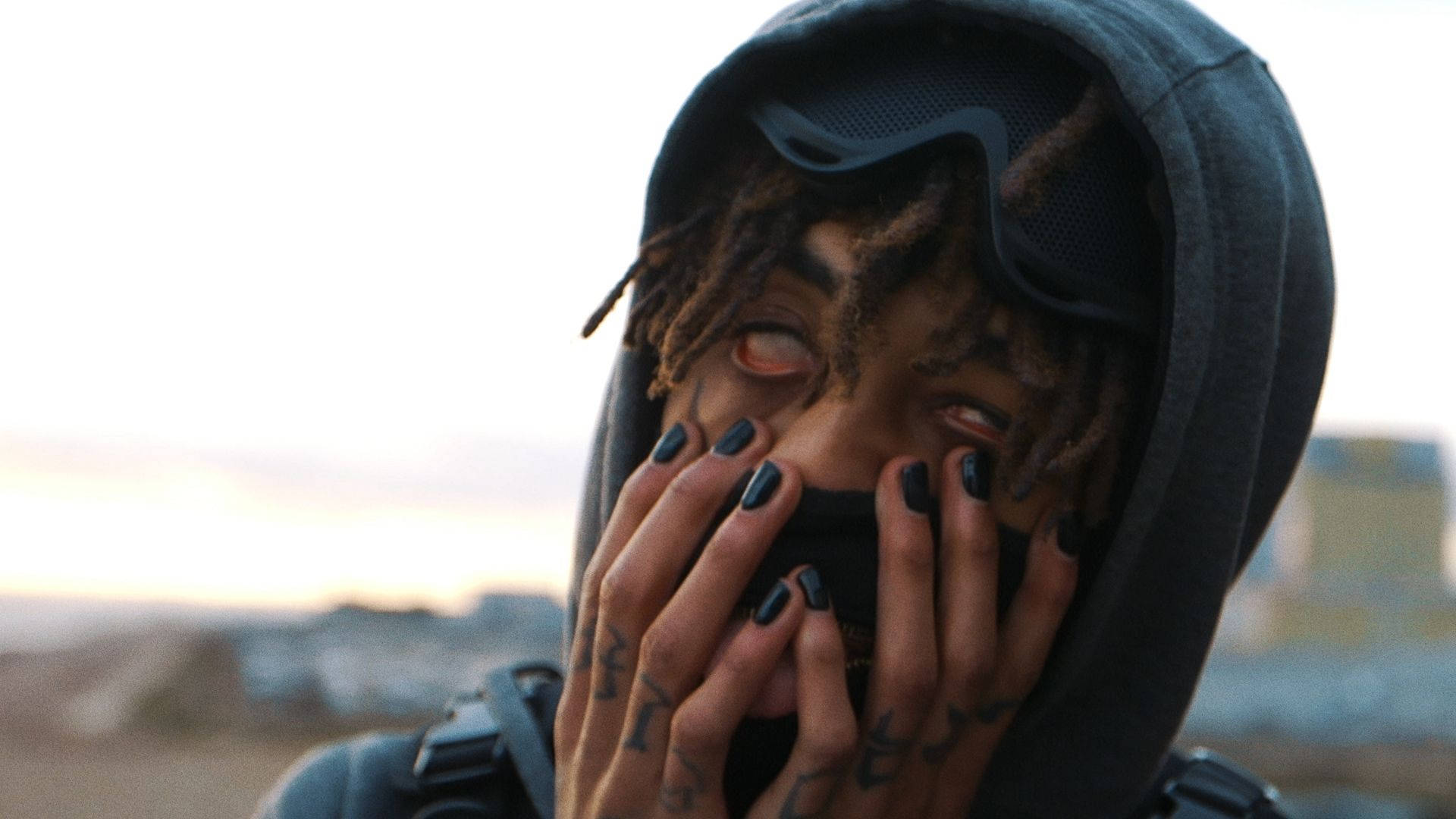 Scarlxrd 桌布 壁紙 — 一個留著辮子、塗黑色指甲油的人，用刺青的手遮住嘴巴，穿著連帽衫和護目鏡，背景是朦朧的城市景觀。