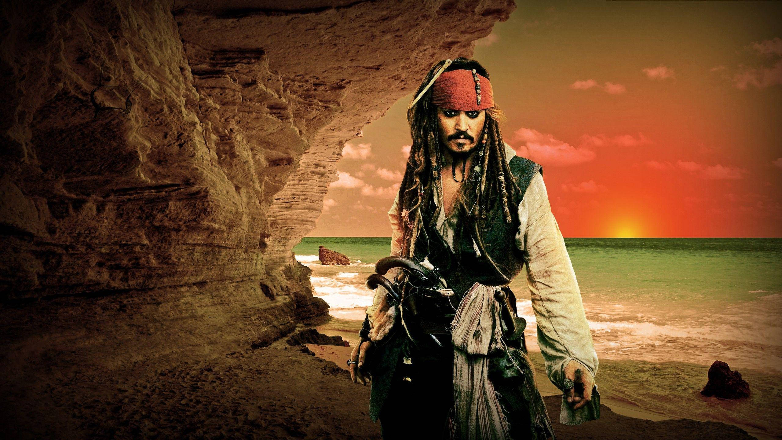 Pirates Of The Caribbean 桌布 壁紙 — 一個留著辮子、戴著紅色頭巾的海盜站在岩石海灘洞穴入口處，熾熱的夕陽在海面上閃耀。