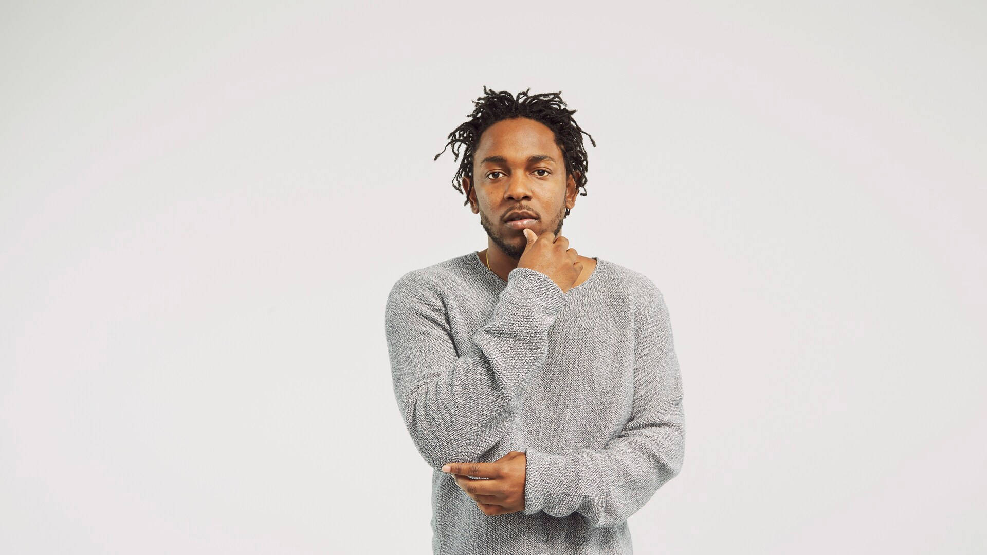 Kendrick 桌布 壁紙 — 一個留著辮子、身穿灰色毛衣的男人在純白色背景前沉思。