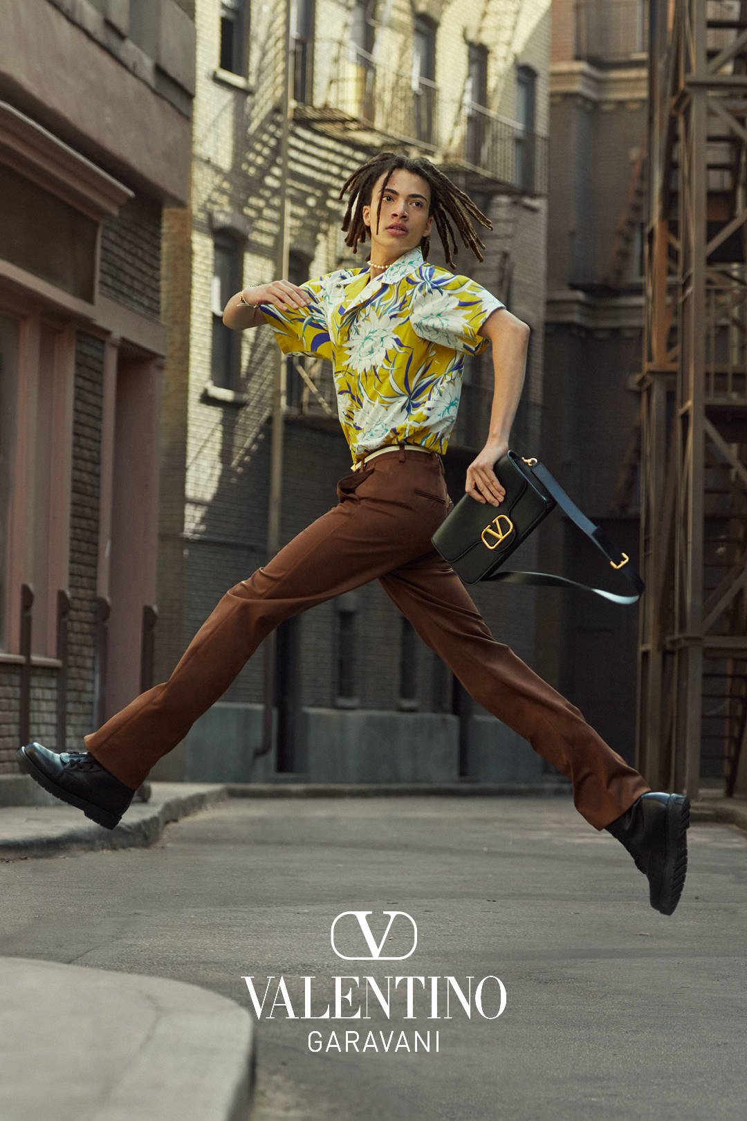Photoshoot 桌布 壁紙 — 一個留著辮子的人穿著鮮豔的花襯衫和棕色褲子，在空中跳躍，手持黑色手提包，背景是粗糙的城市小巷。
