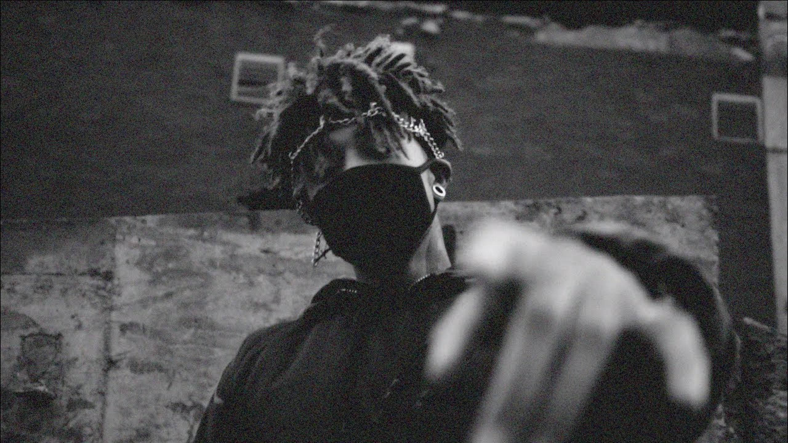 Scarlxrd 桌布 壁紙 — 一個留著辮子頭、戴著口罩的人，在粗糙的城市牆壁背景下，向觀眾伸出一隻模糊的手。