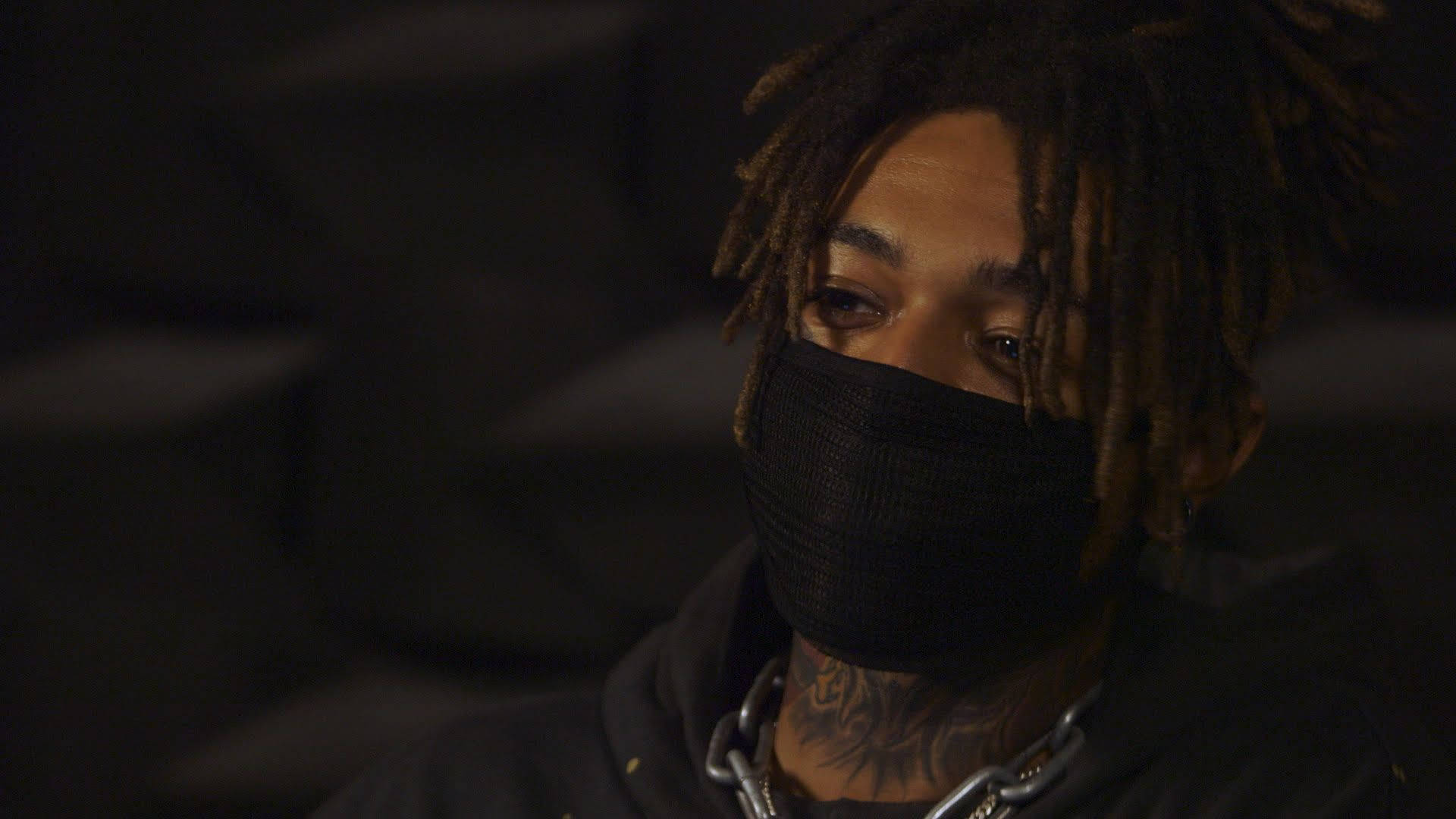 scarlxrd 桌布 壁紙 — 一個留著辮子頭、戴黑色口罩的人，在昏暗燈光下若有所思地凝視，可見刺青和鏈條項鍊。