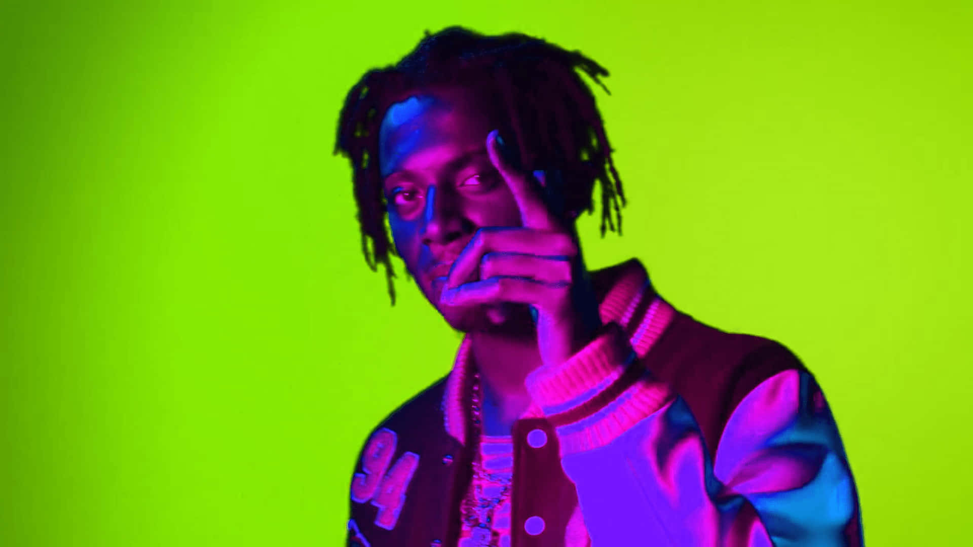 Playboi Carti 桌布 壁紙 — 一個留著辮子頭、身穿大學夾克的人，在紫色和藍色燈光下，以螢光綠色背景前擺姿勢。