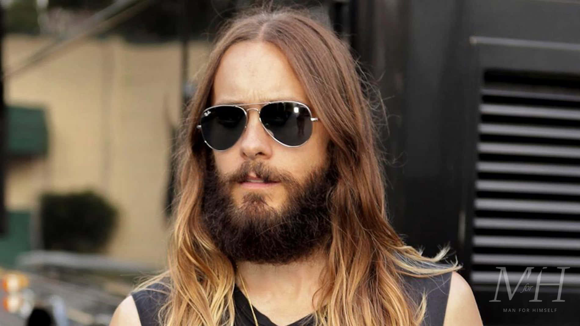 jared leto 桌布 壁紙 — 一個留著長棕色頭髮和滿臉鬍鬚的男人，在城市環境中戴著飛行員太陽眼鏡。