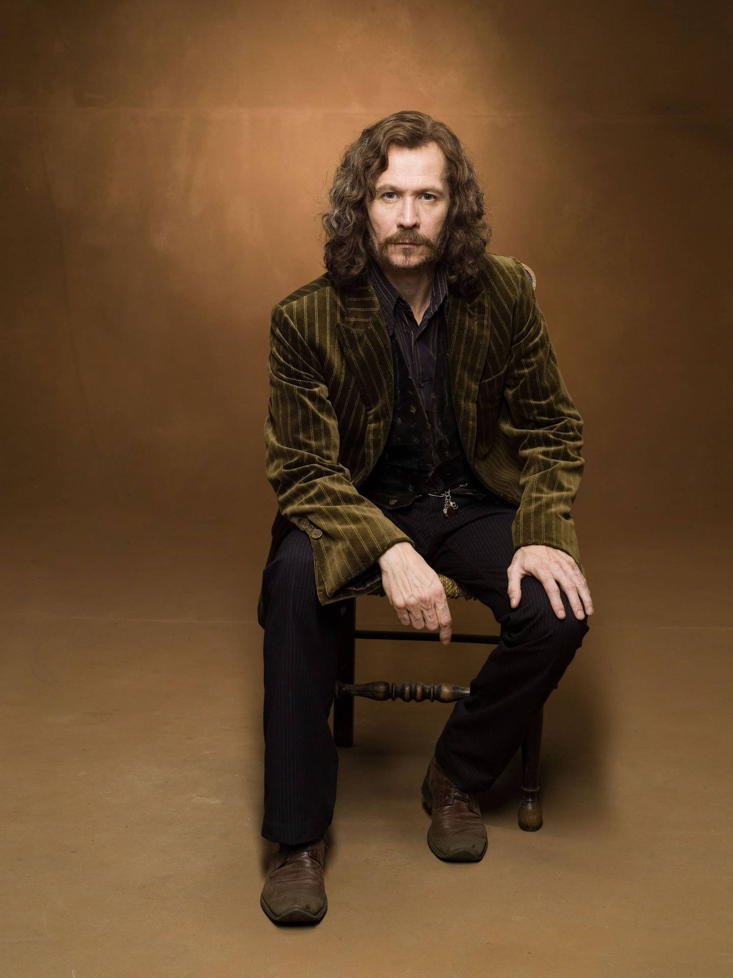 Sirius Black 桌布 壁紙 — 一個留著長波浪頭髮和鬍鬚的男人坐在木椅上，穿著綠色細條紋天鵝絨外套，背景是溫暖的棕色攝影棚。