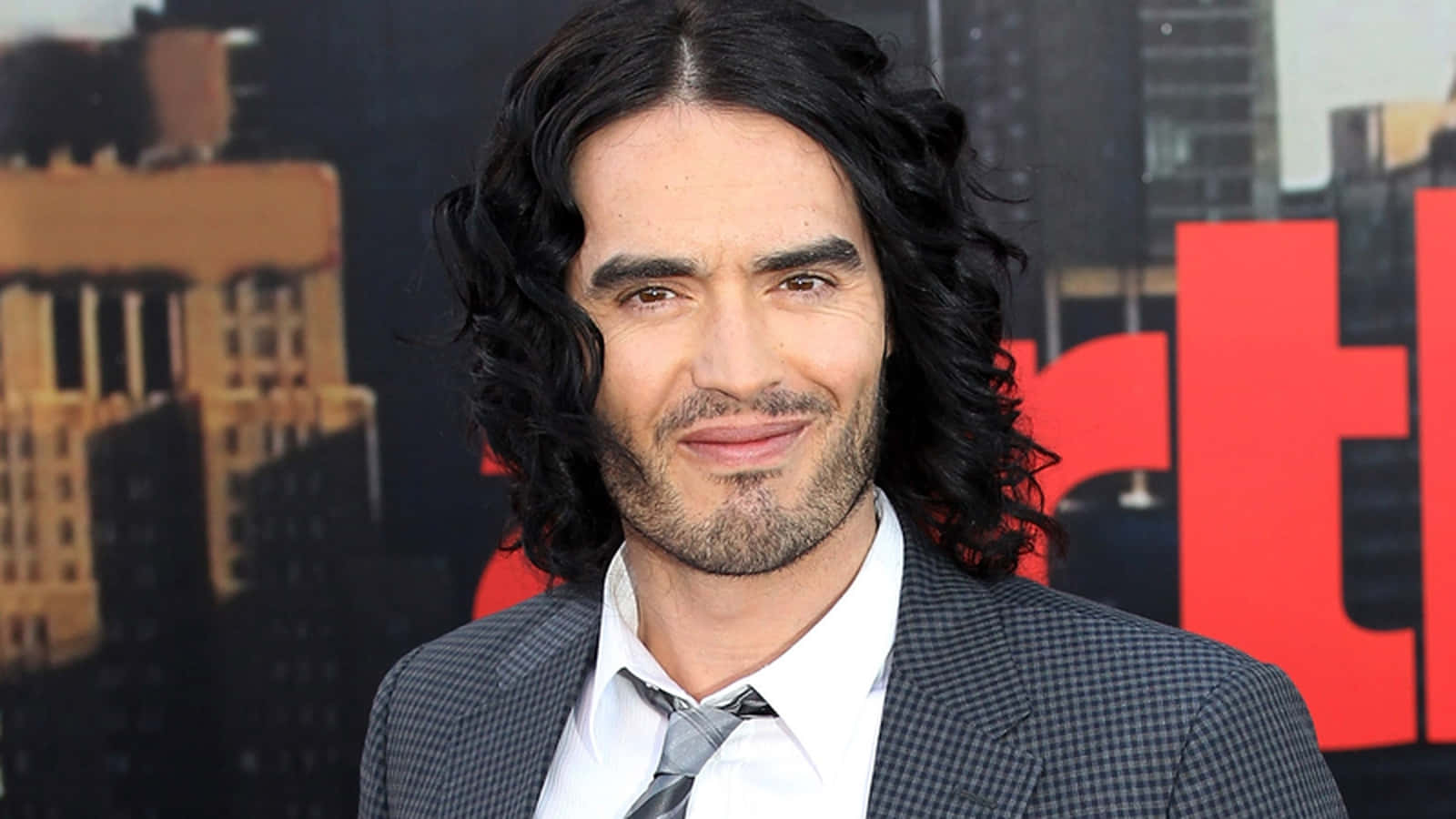 Celebrity 桌布 壁紙 — 一個留著長深色捲髮和鬍鬚的男人，穿著格紋西裝，在模糊的城市背景前微笑，背景有紅色字樣。