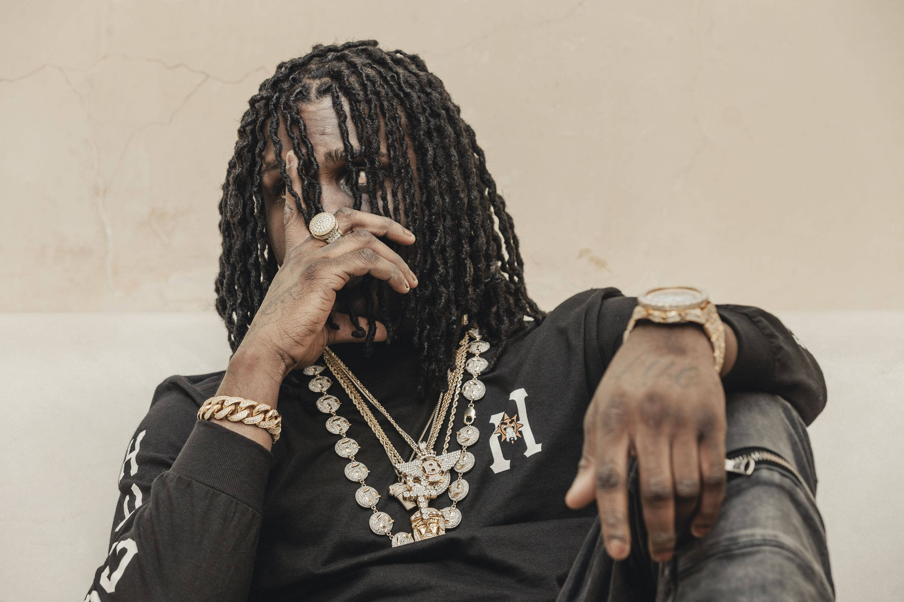 Chief Keef 桌布 壁紙 — 一個留著長辮子、戴著金飾的男人，靠著質感米色牆壁沉思，手半掩著臉。