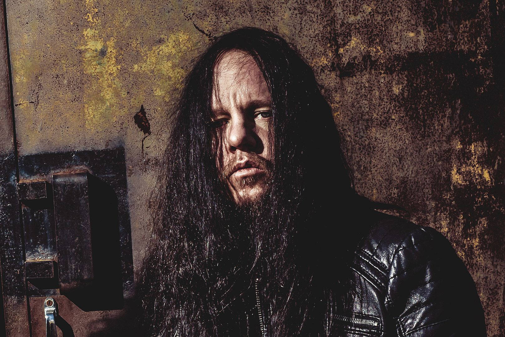 Joey Jordison 桌布 壁紙 — 一個留著長黑髮和鬍鬚的男人，穿著黑色皮夾克，背景是生鏽的金屬牆，光線充滿戲劇性。