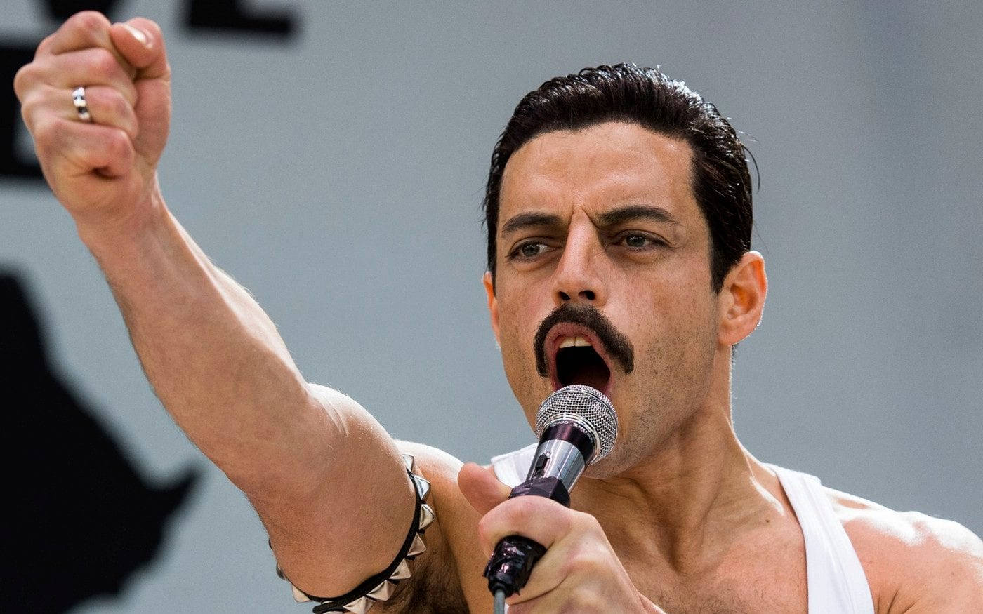 Rami Malek 桌布 壁紙 — 一個留著鬍子的男人，穿著白色背心，一邊舉起拳頭，一邊熱情地對著麥克風唱歌。