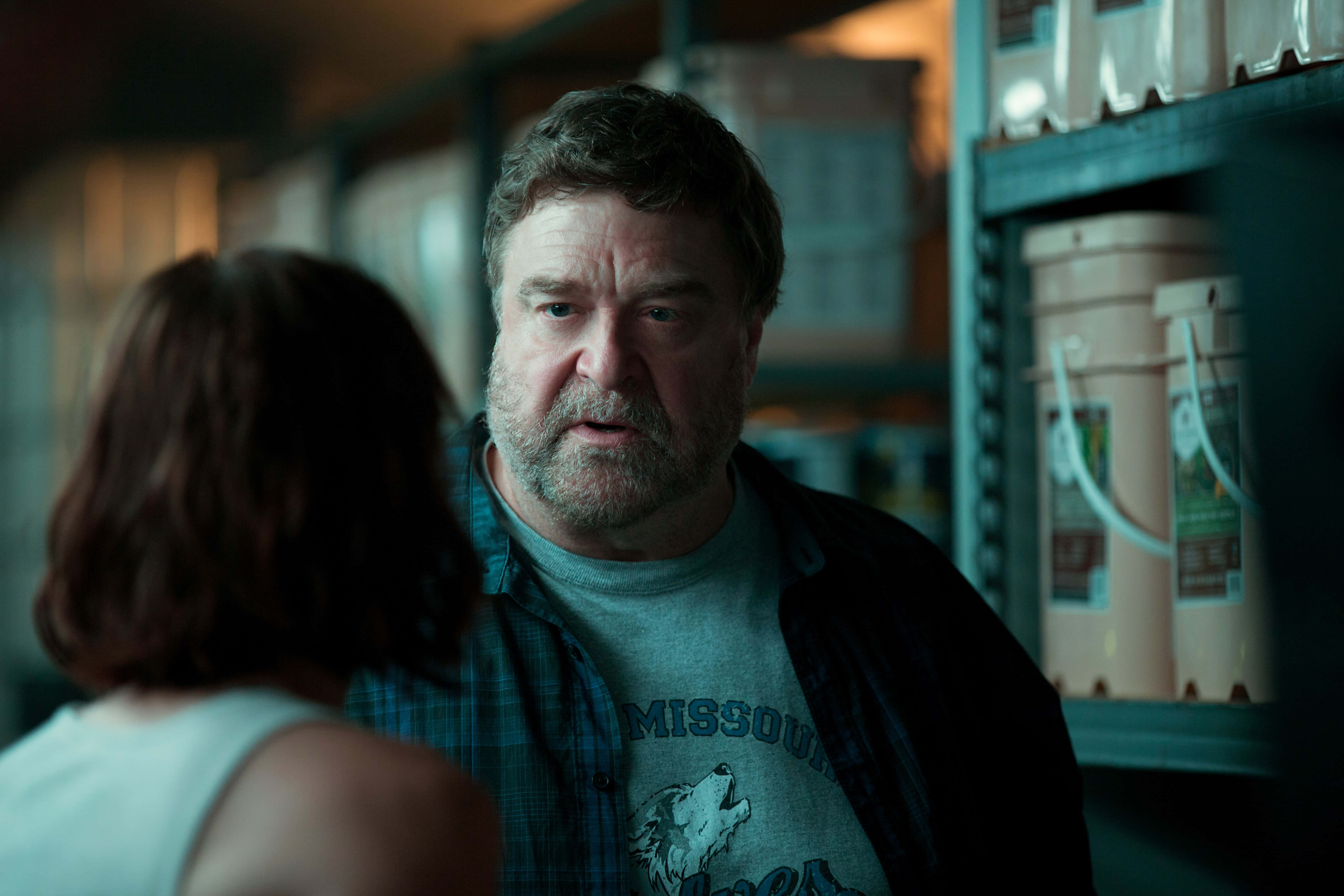 John Goodman 桌布 壁紙 — 一個留著鬍鬚、穿著格紋襯衫的男人在燈光昏暗的倉庫通道中專注地凝視著一個女人。