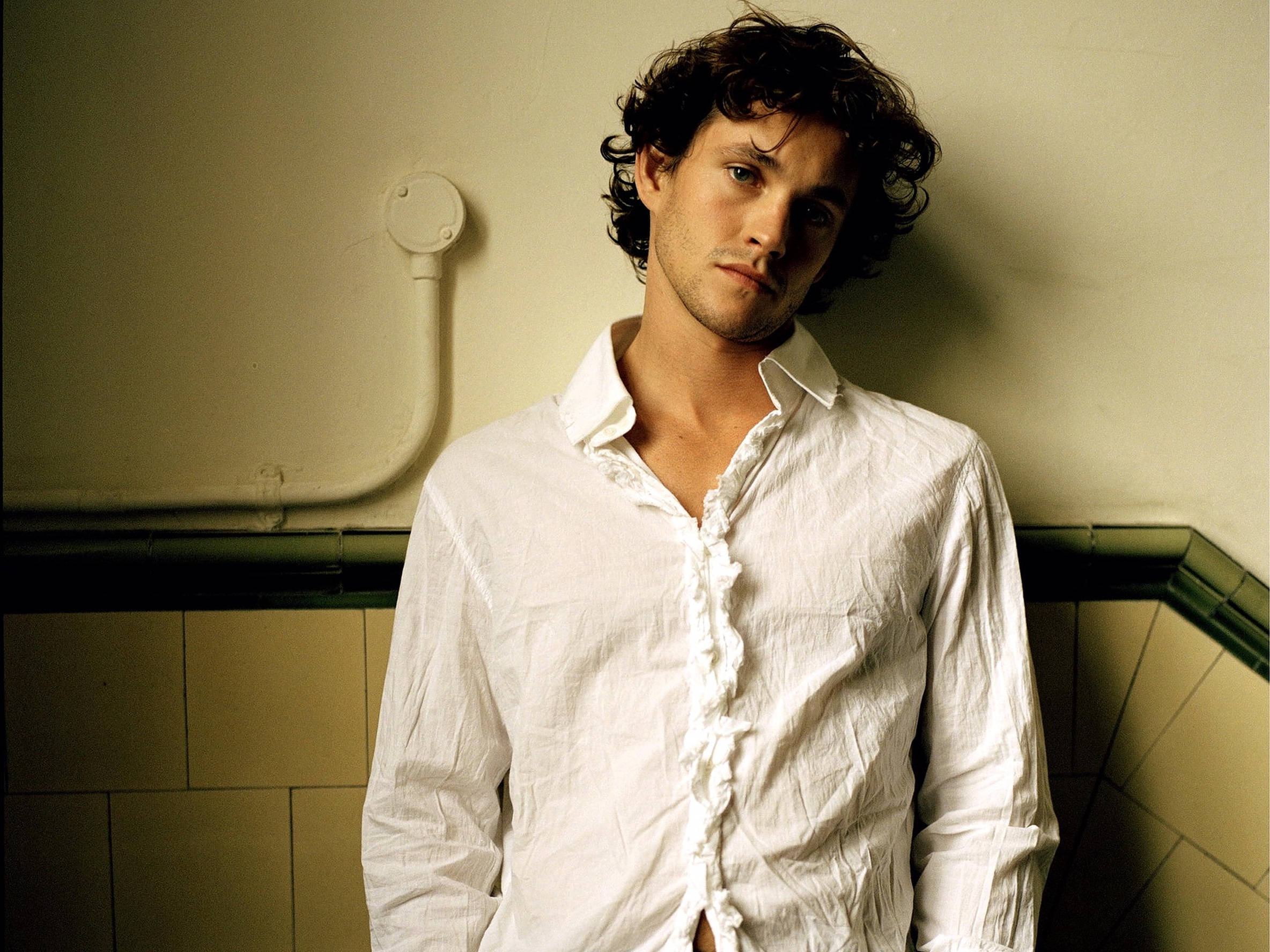 Hugh Dancy 桌布 壁紙 — 一個留著黑色捲髮的男人，在溫暖的燈光下，穿著皺巴巴的白襯衫，靠在瓷磚牆上。