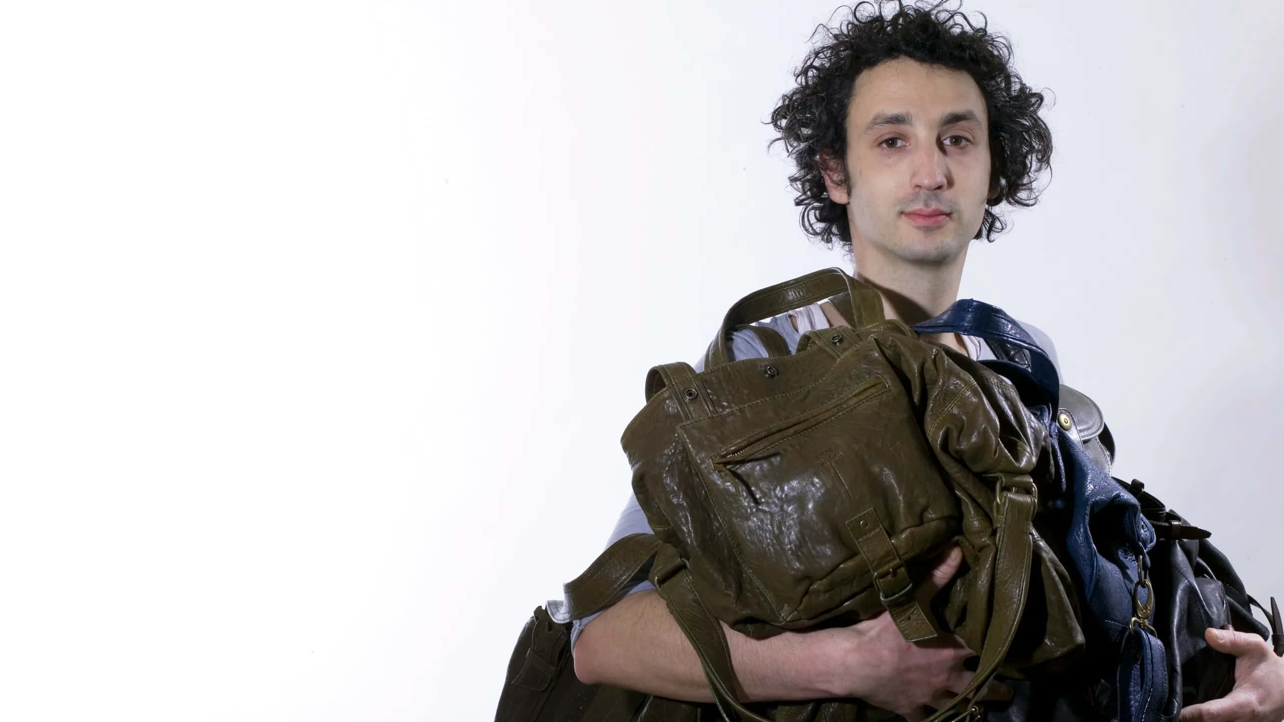 Handbag 桌布 壁紙 — 一個留著黑色捲髮的男人，在純白背景前拿著多個磨損的皮革袋。