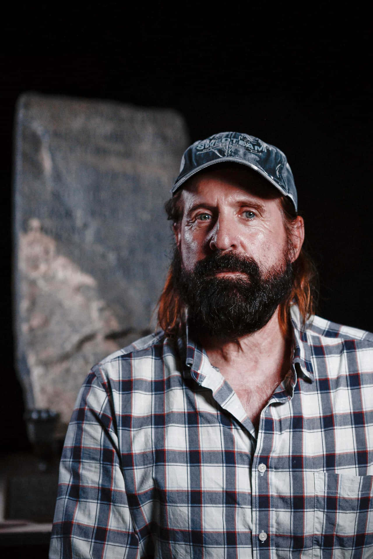 Peter Stormare 桌布 壁紙 — 一個留長鬍鬚的男人，長髮，穿著格紋襯衫和帽子，背景黑暗，身後有一塊石板。