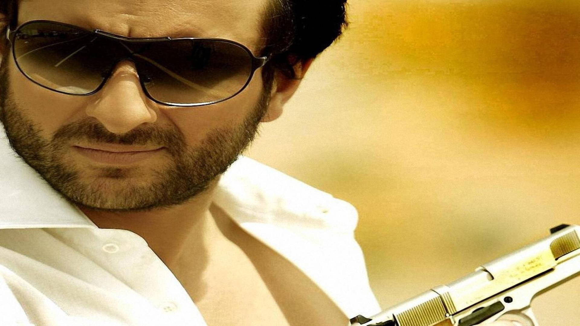 Saif Ali Khan 桌布 壁紙 — 一個留鬍子、戴太陽眼鏡的男人，在溫暖的金色背景前拿著一把銀色手槍。