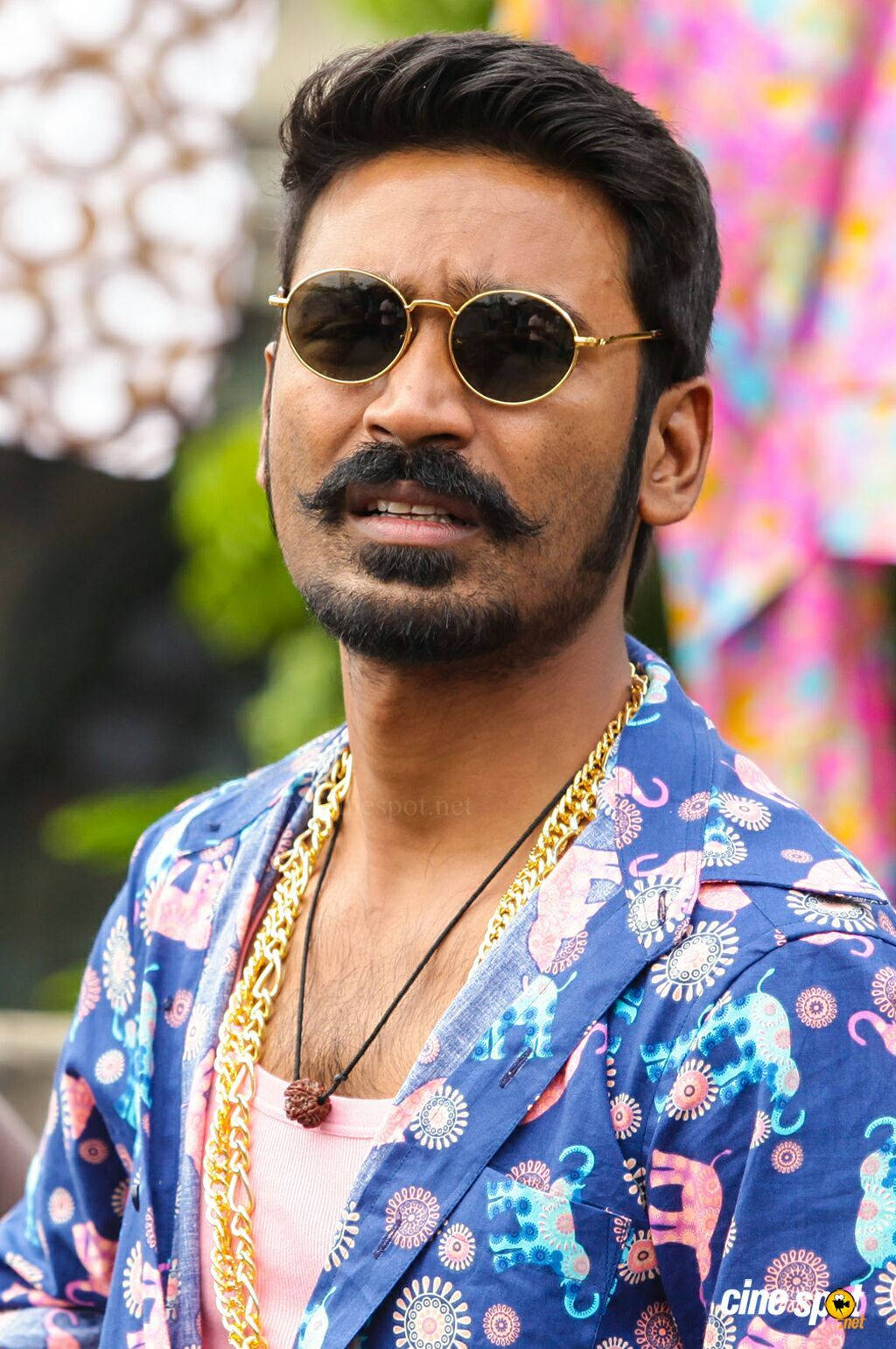 Indian 桌布 壁紙 — 一個留鬍子、戴太陽眼鏡的男人，穿著鮮豔的藍色圖案外套和金色鏈條，背景是色彩繽紛的模糊效果。