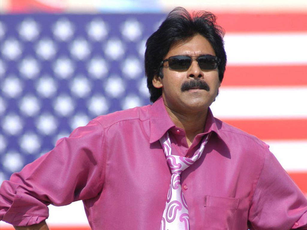 Pawan Kalyan 桌布 壁紙 — 一個留鬍子、戴太陽眼鏡的男人自信地站在模糊的美國國旗前，身穿粉紅色襯衫和圖案領帶。