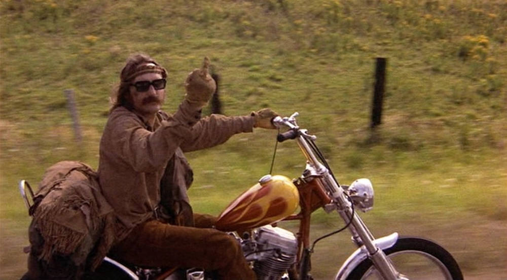 Easy Rider 桌布 壁紙 — 一個留鬍子、戴太陽眼鏡的男人騎著火焰圖案的機車穿過草地，豎起大拇指。