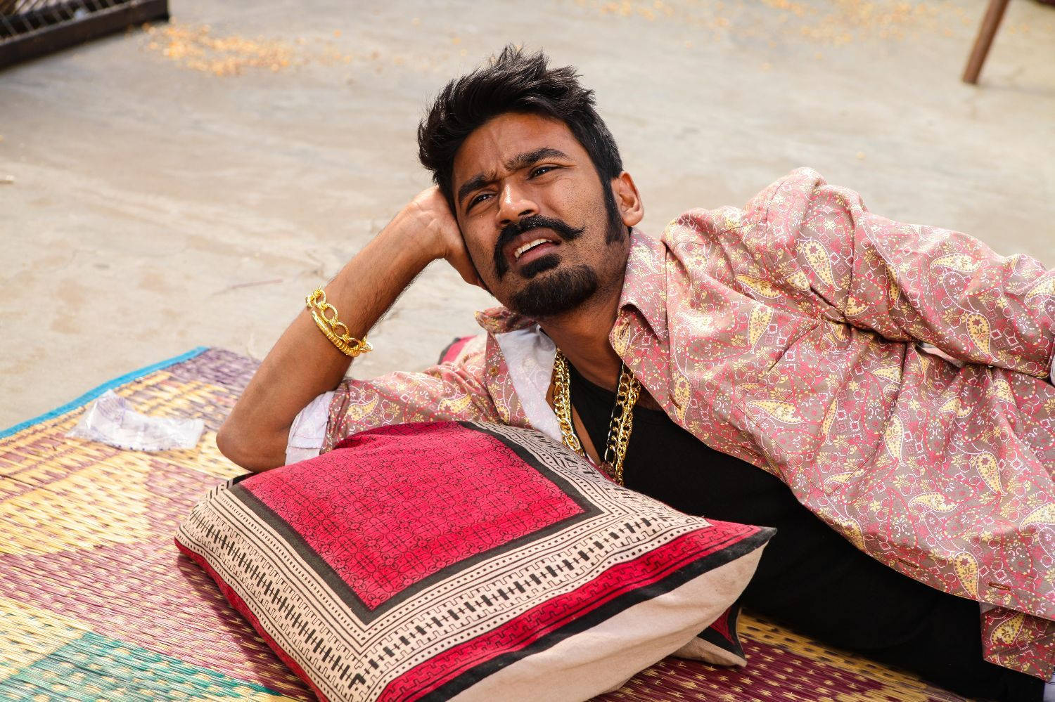 Dhanush 桌布 壁紙 — 一個留鬍子、戴金飾的男人躺在色彩繽紛的圖案地毯上，頭枕在紅色枕頭上休息。
