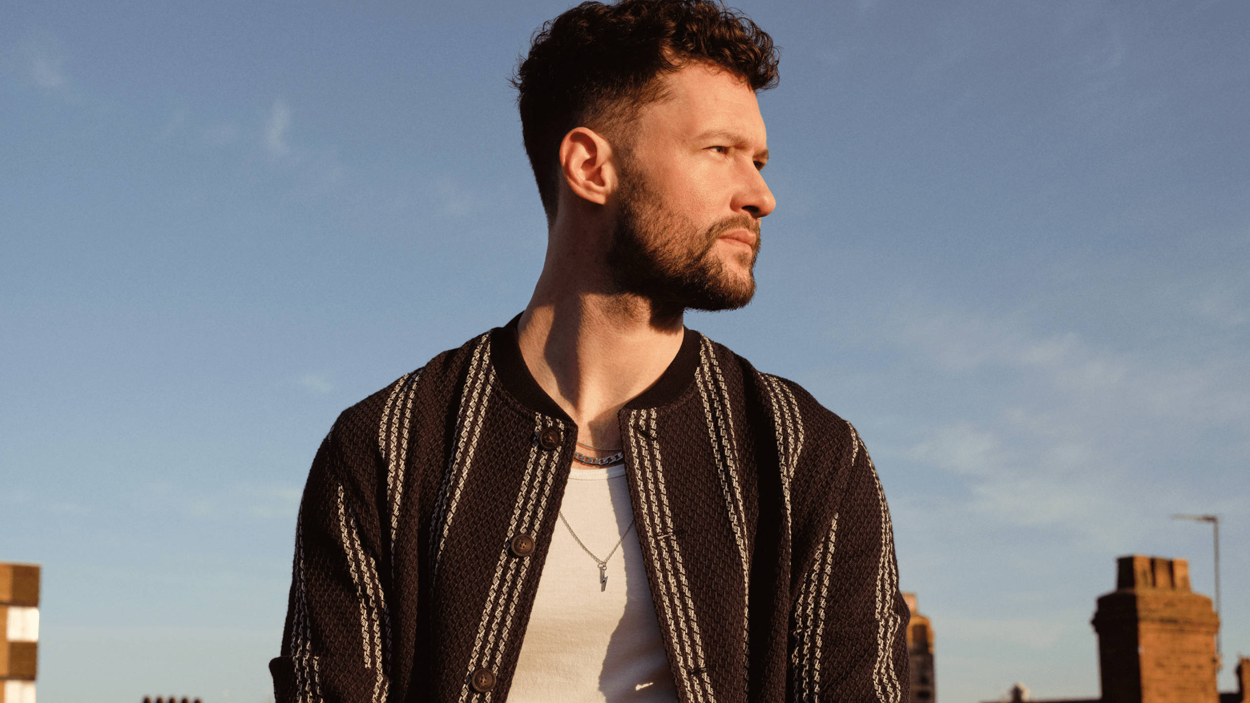 Calum Scott 桌布 壁紙 — 一個留鬍子、捲髮的男人側身站在清澈的藍天和城市屋頂背景下。
