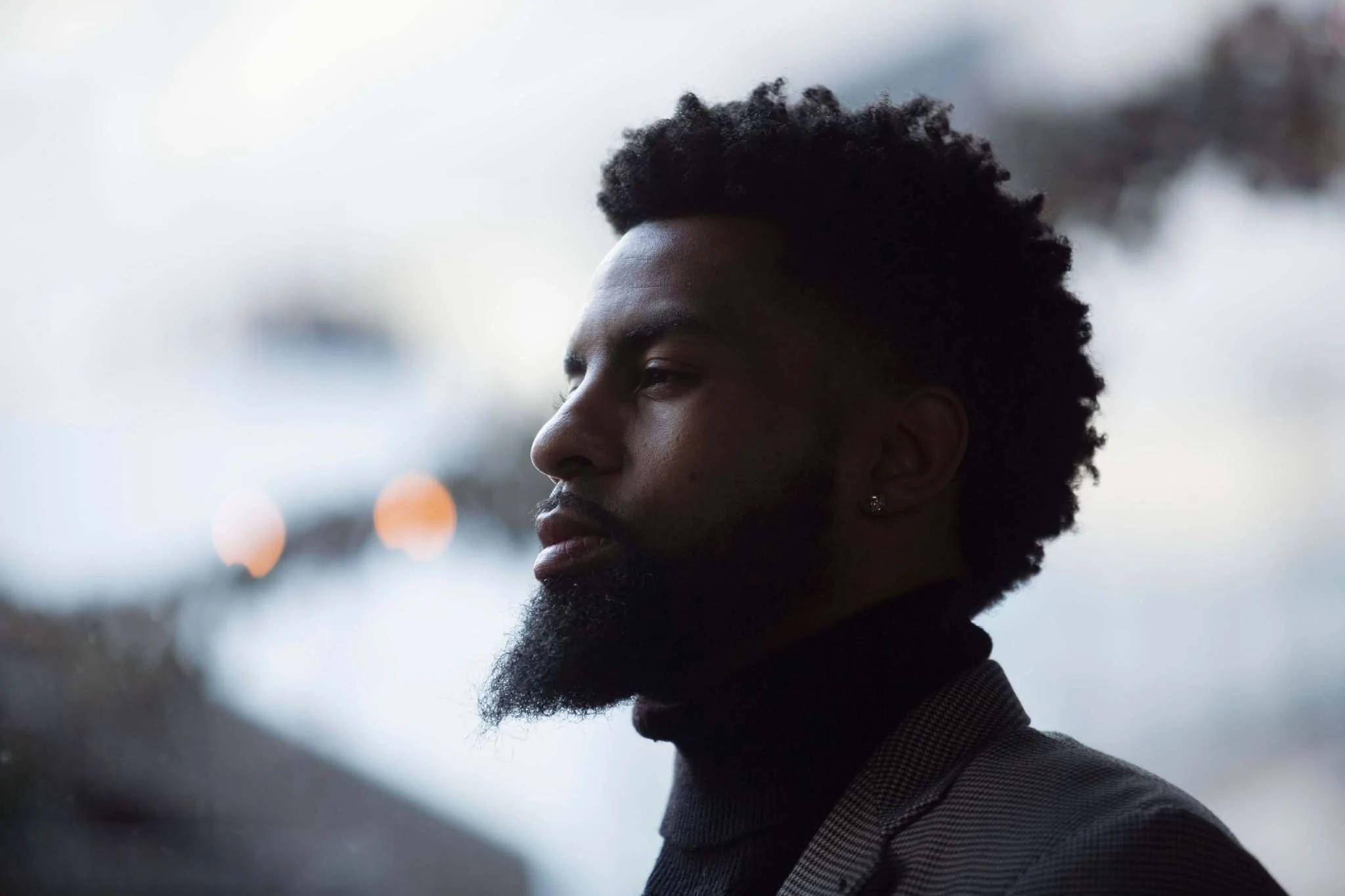 Black Man 桌布 壁紙 — 一個留鬍子、梳非洲式髮型的男人若有所思地望向窗外，背景是模糊的城市燈光。