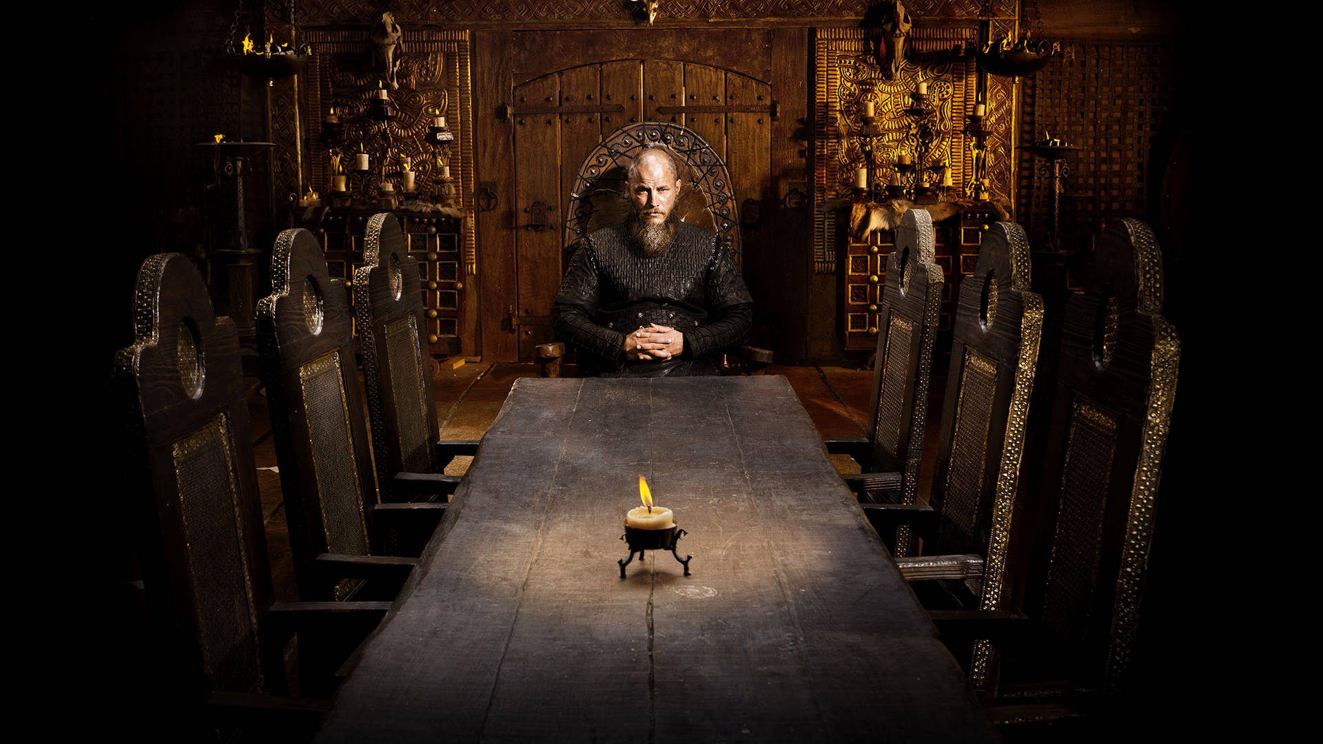 Vikings 桌布 壁紙 — 一個留鬍子、身穿盔甲的男人莊嚴地坐在王座上，身處一個昏暗的中世紀大廳，僅由長桌上的一根蠟燭照亮。