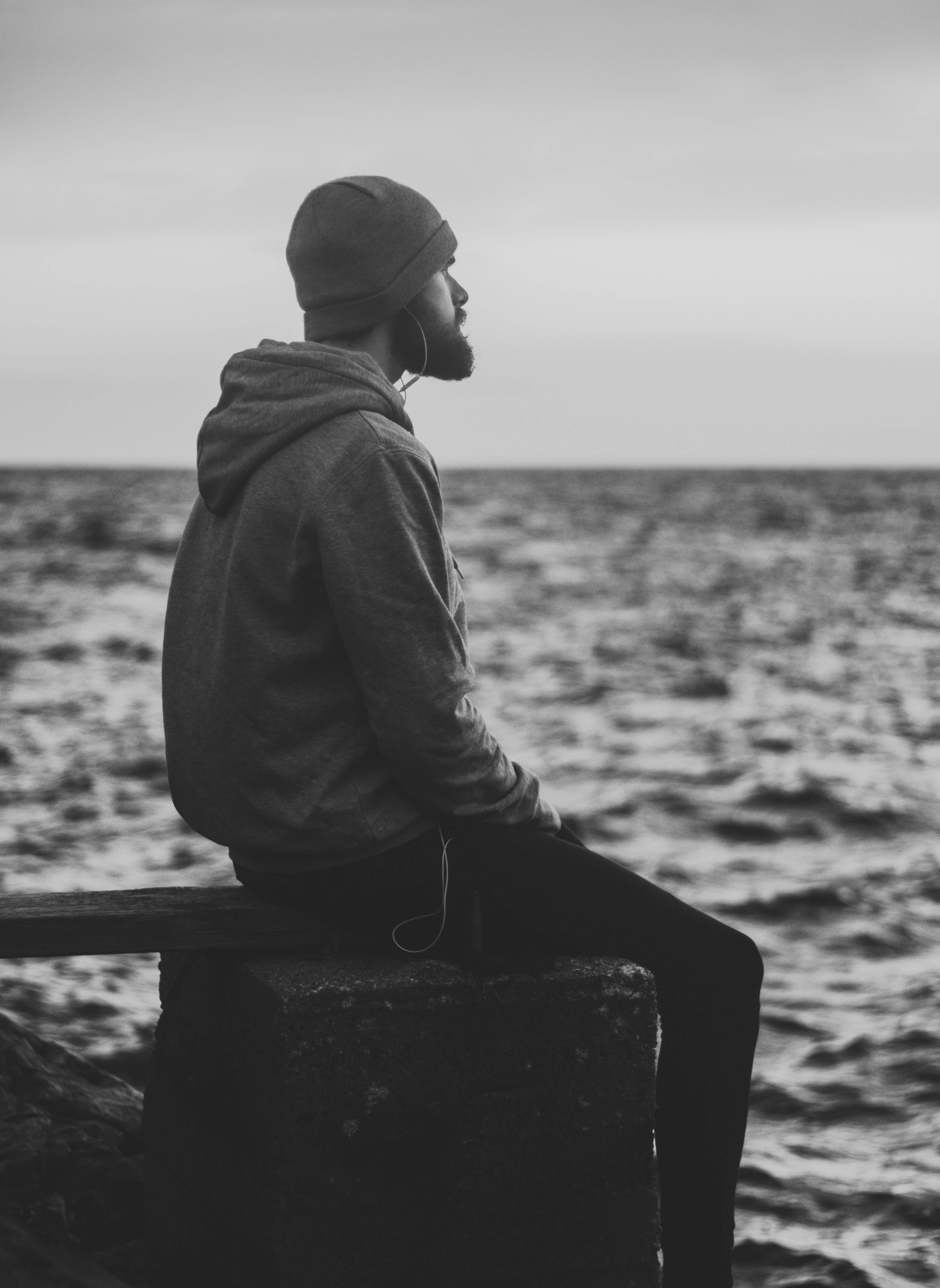 Alone 桌布 壁紙 — 一個留鬍子戴毛帽的男人獨自坐在石碼頭上，凝視著陰天下一望無際的海洋。