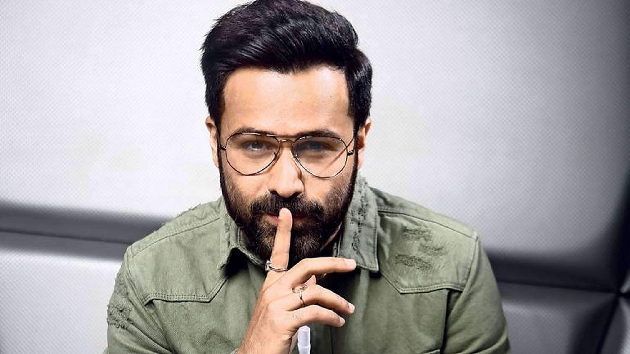 Emraan Hashmi 桌布 壁紙 — 一個留鬍子戴眼鏡的男人，在紋理灰色背景前將手指放在嘴唇上做出安靜的手勢。