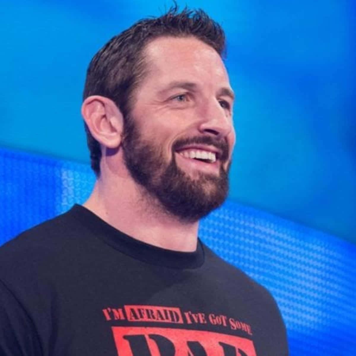 WWE 桌布 壁紙 — 一個留鬍子的微笑男子，身穿印有紅色文字的黑色T恤，背景為藍色數位背景。