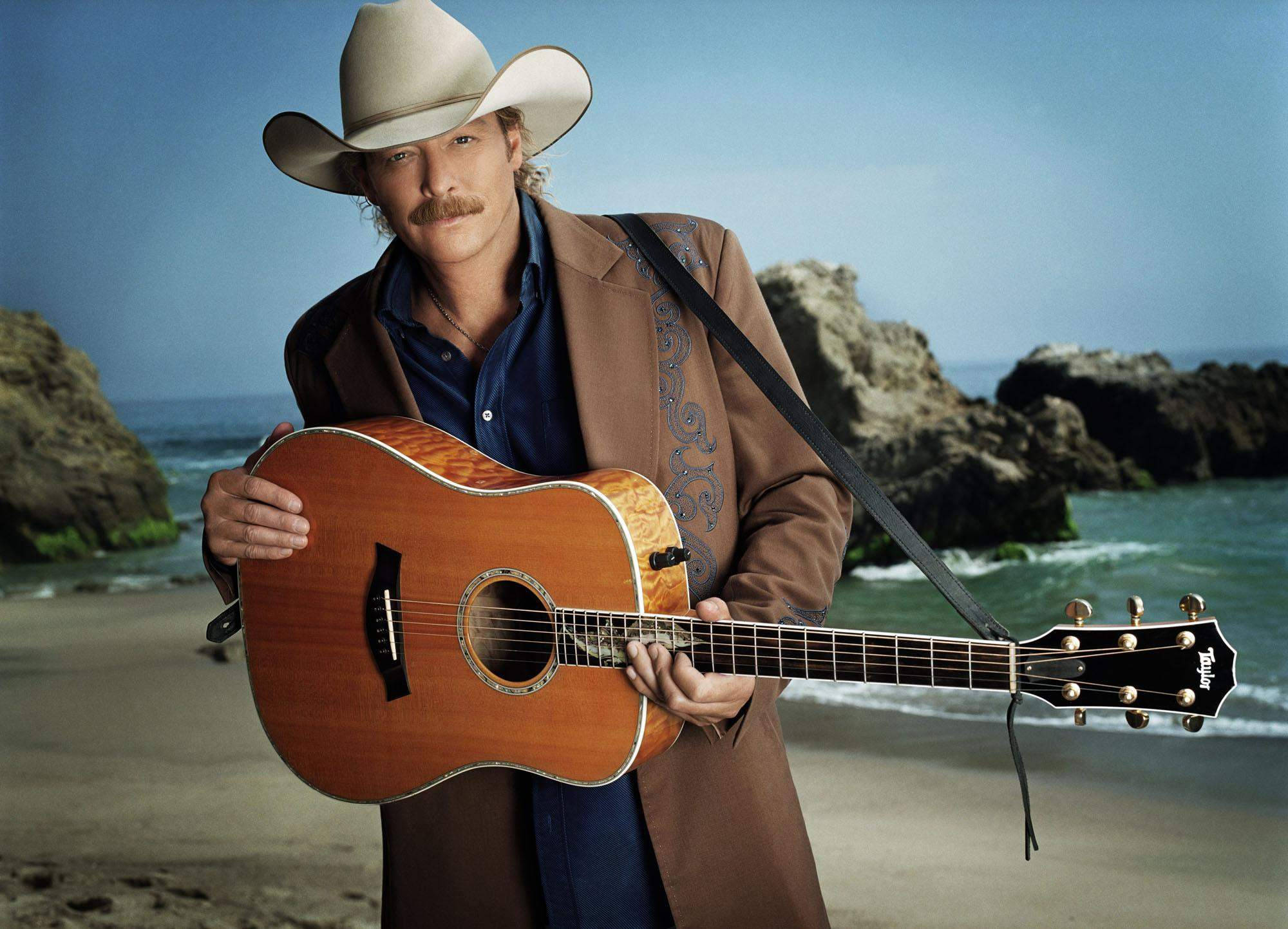 Alan Jackson 桌布 壁紙 — 一個留鬍子的牛仔在岩石海灘上抱著一把原聲吉他，身後海浪拍打。