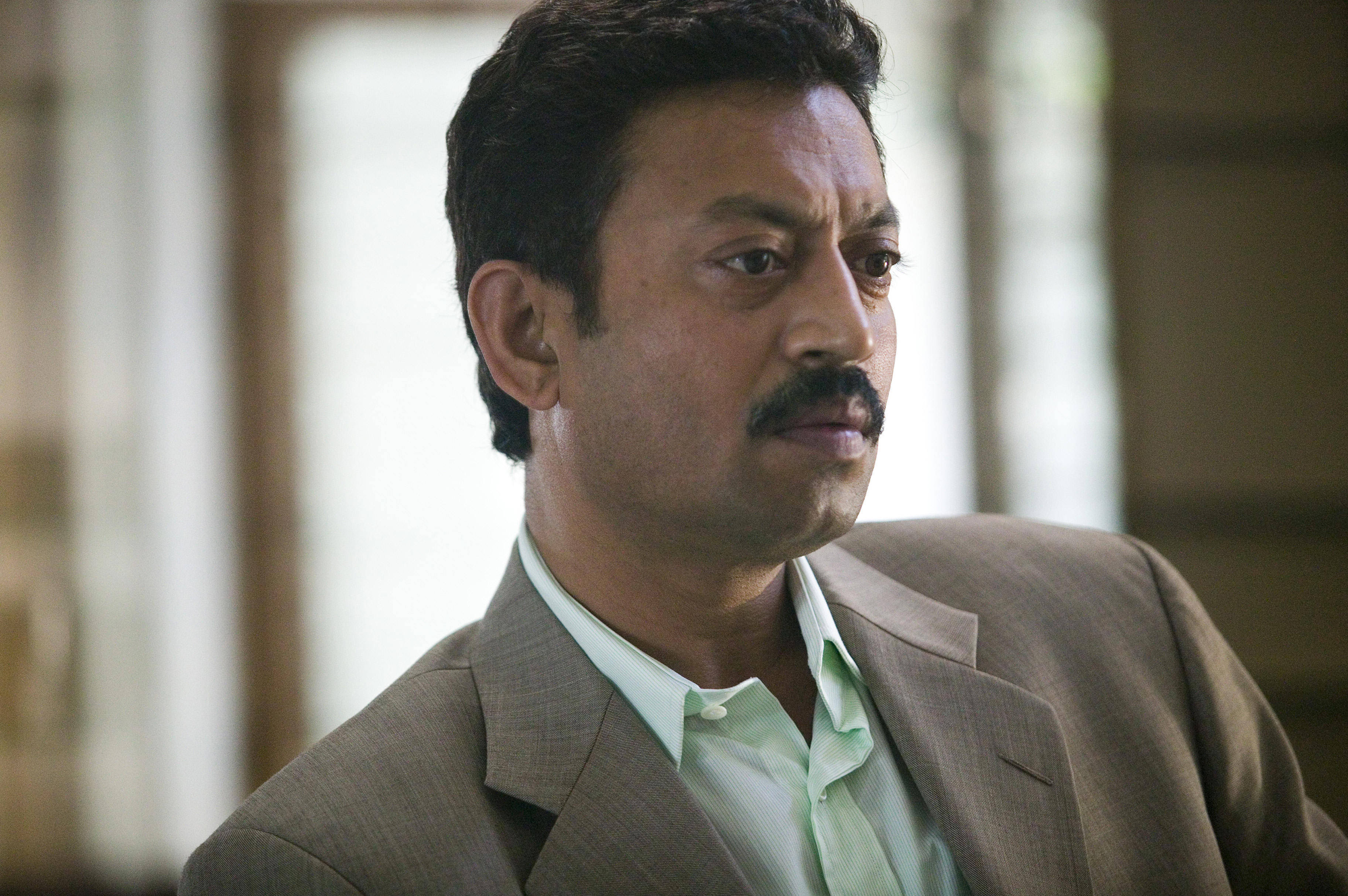 Irrfan Khan 桌布 壁紙 — 一個留鬍子的男人，穿著棕色西裝和淺綠色襯衫，表情嚴肅地看著鏡頭外。