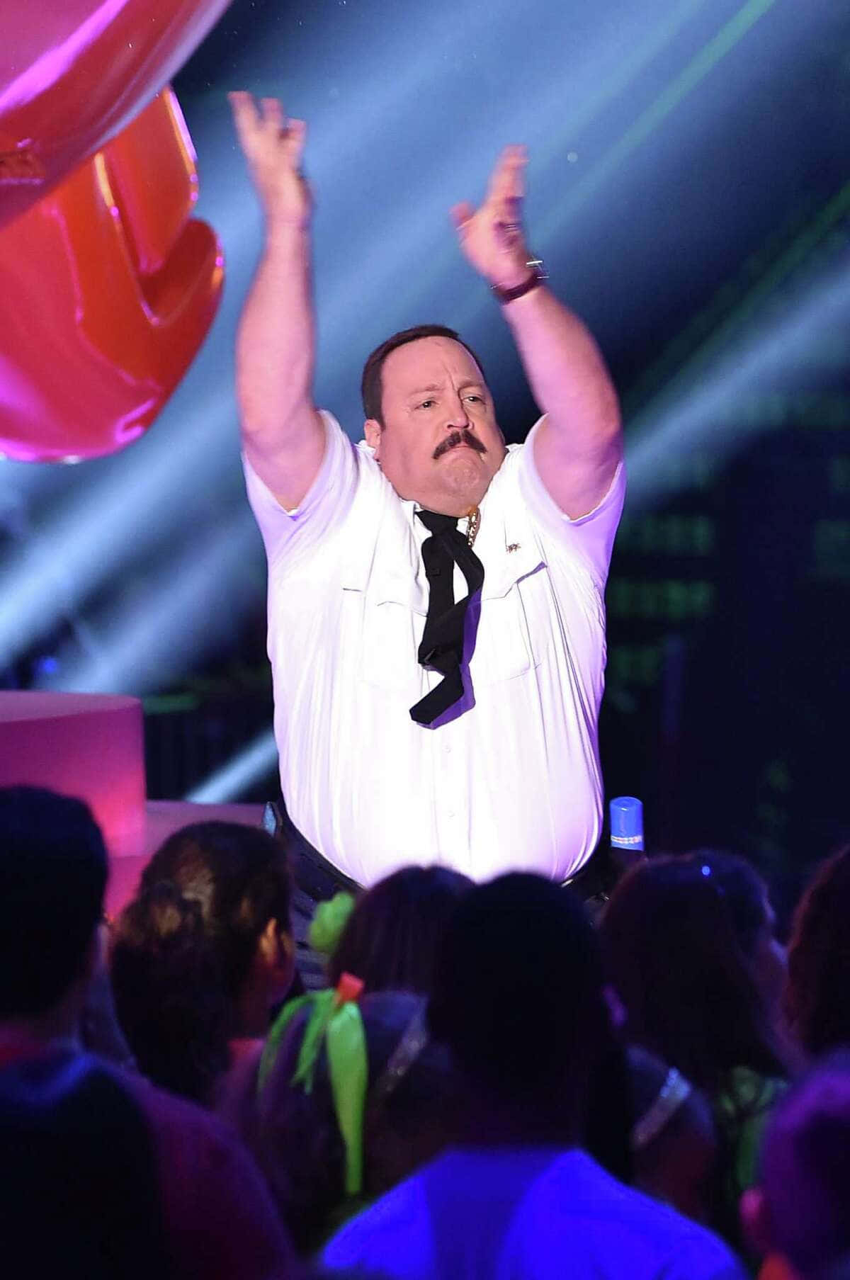 Kevin James 桌布 壁紙 — 一個留鬍子的男人，穿著白襯衫和黑色領帶，在一個燈光明亮、有彩色聚光燈的舞台上舉起雙臂，台下有群眾。