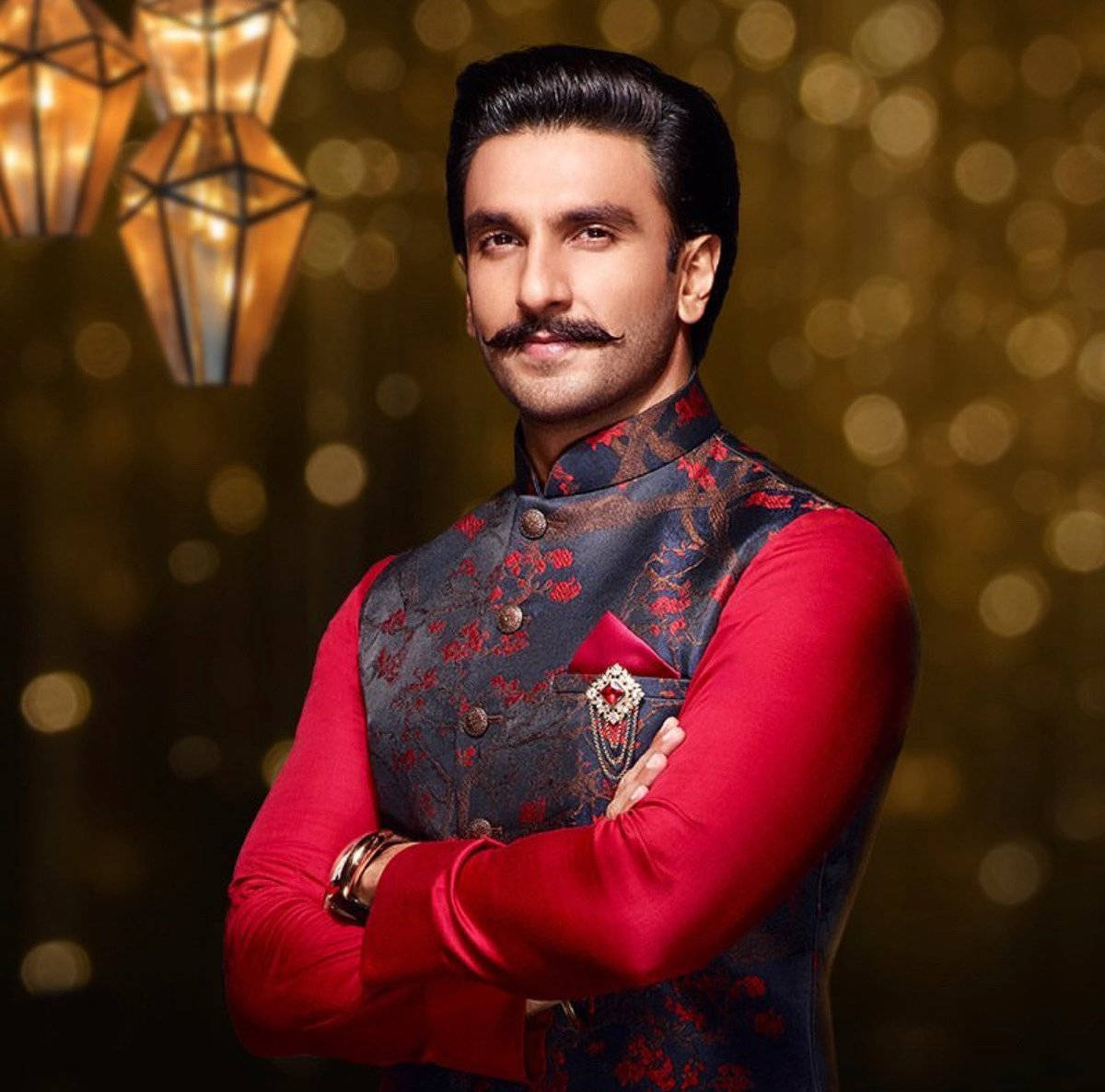 Ranveer Singh 桌布 壁紙 — 一個留鬍子的男人，身穿紅色圖案背心，雙臂交叉，自信地站在金色散景背景前，背景有懸掛的燈籠。