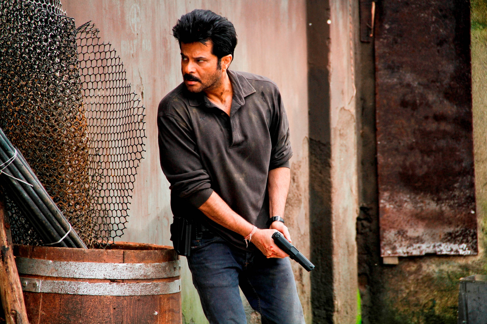 anil kapoor 桌布 壁紙 — 一個留鬍子的男人在一條骯髒的後巷，在一個生鏽的桶和鐵絲網旁邊拿著一把手槍。