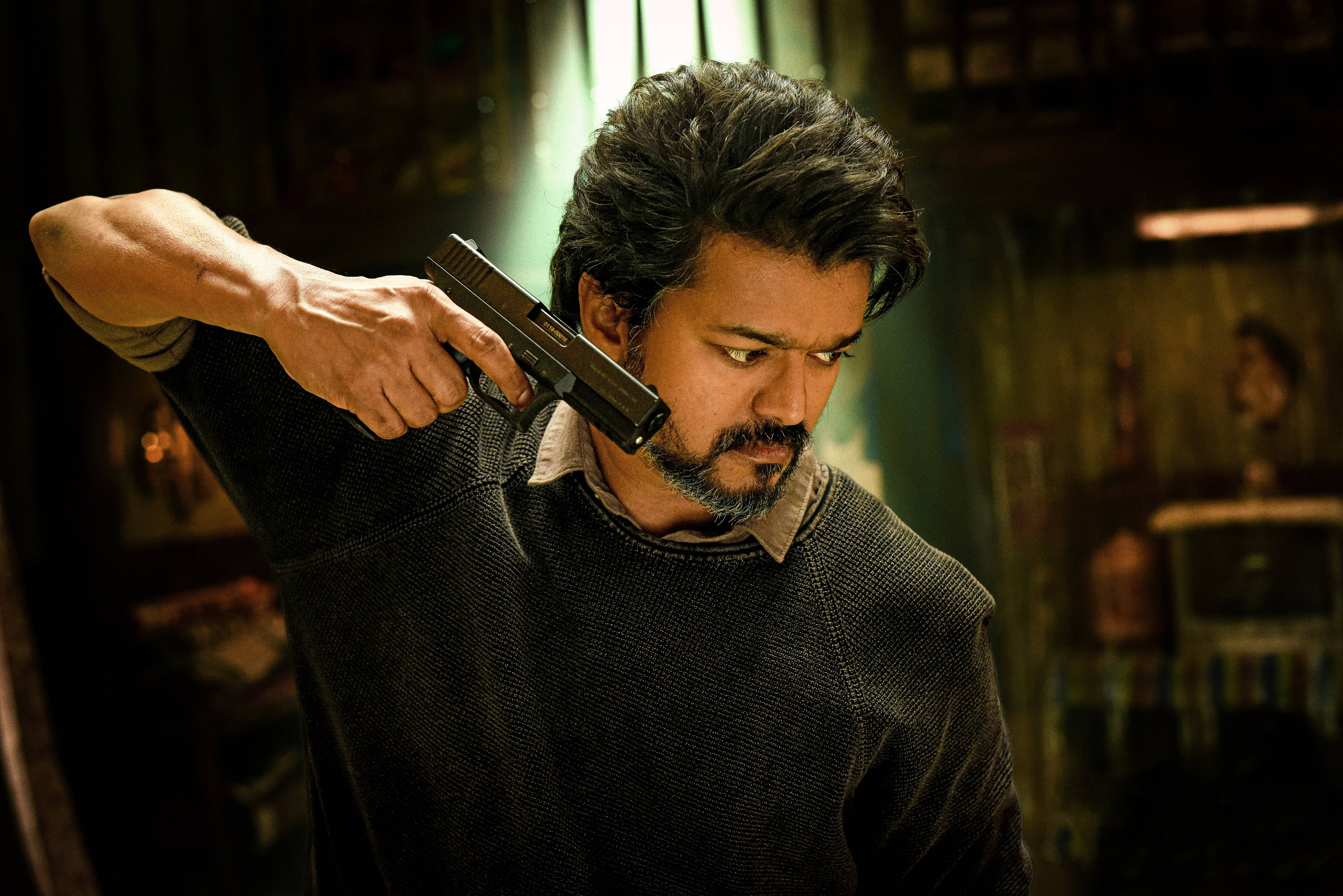 vijay HD 桌布 壁紙 — 一個留鬍子的男人在光線昏暗、有溫暖燈光和木製家具的房間裡，把槍對準自己的太陽穴。