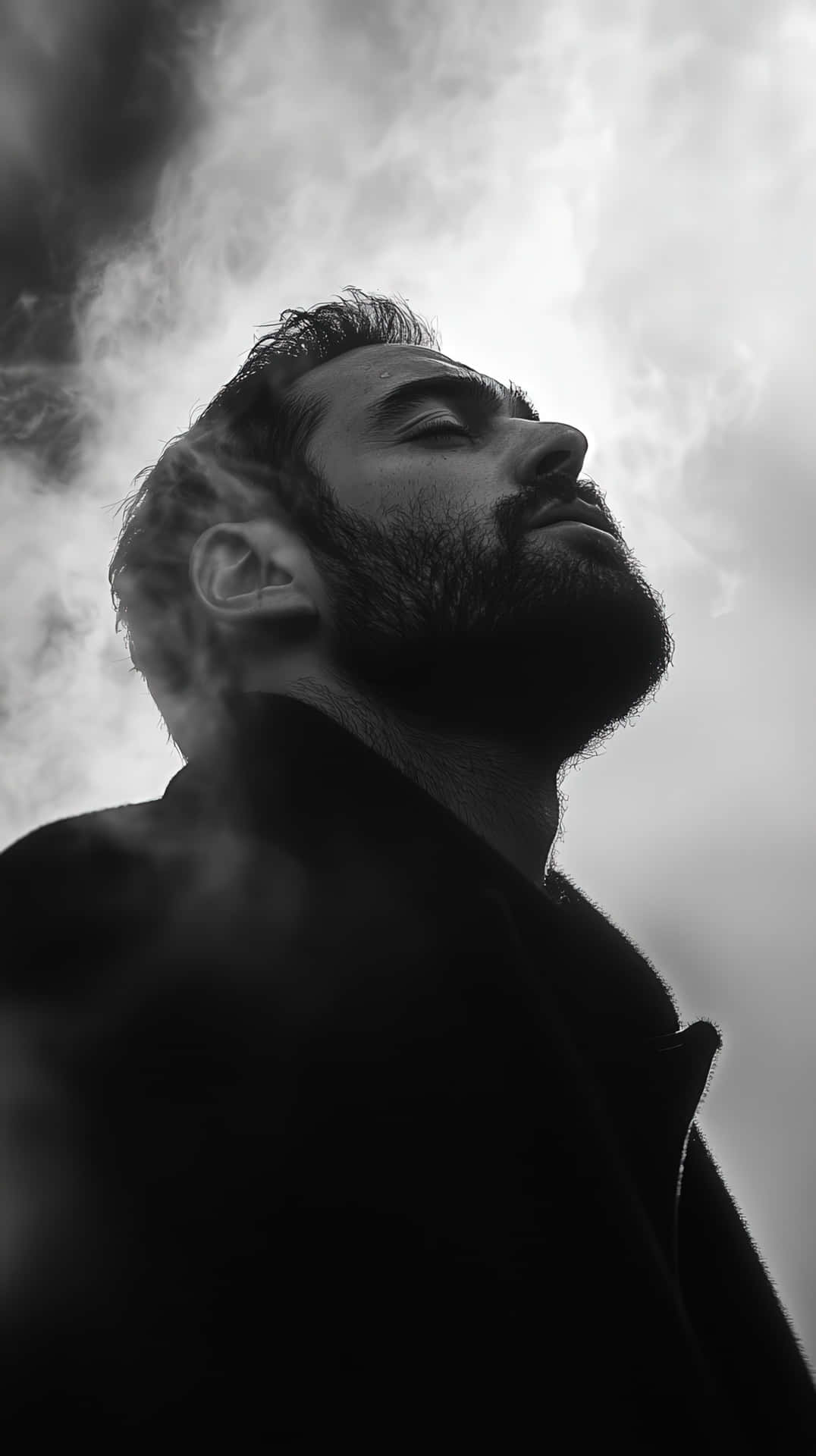 Introspective bearded man in black and white portrait with smoke and dramatic lighting 壁紙 — 一個留鬍子的男人在陰天背景下向上仰望，形成戲劇性的黑白肖像。