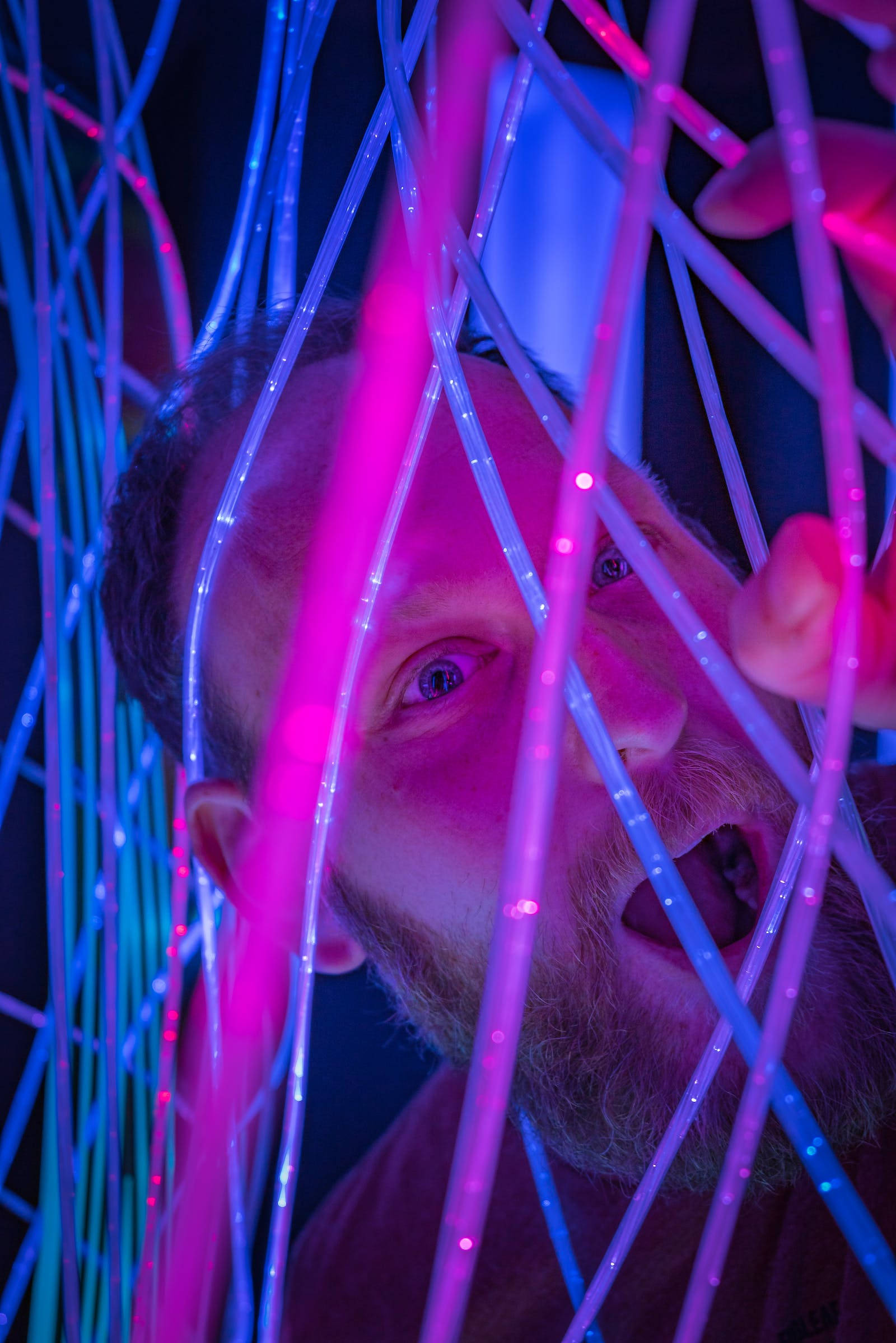 Neon Phone 桌布 壁紙 — 一個留鬍子的男人在黑暗的環境中透過發光的粉紅色和藍色光纖線凝視。