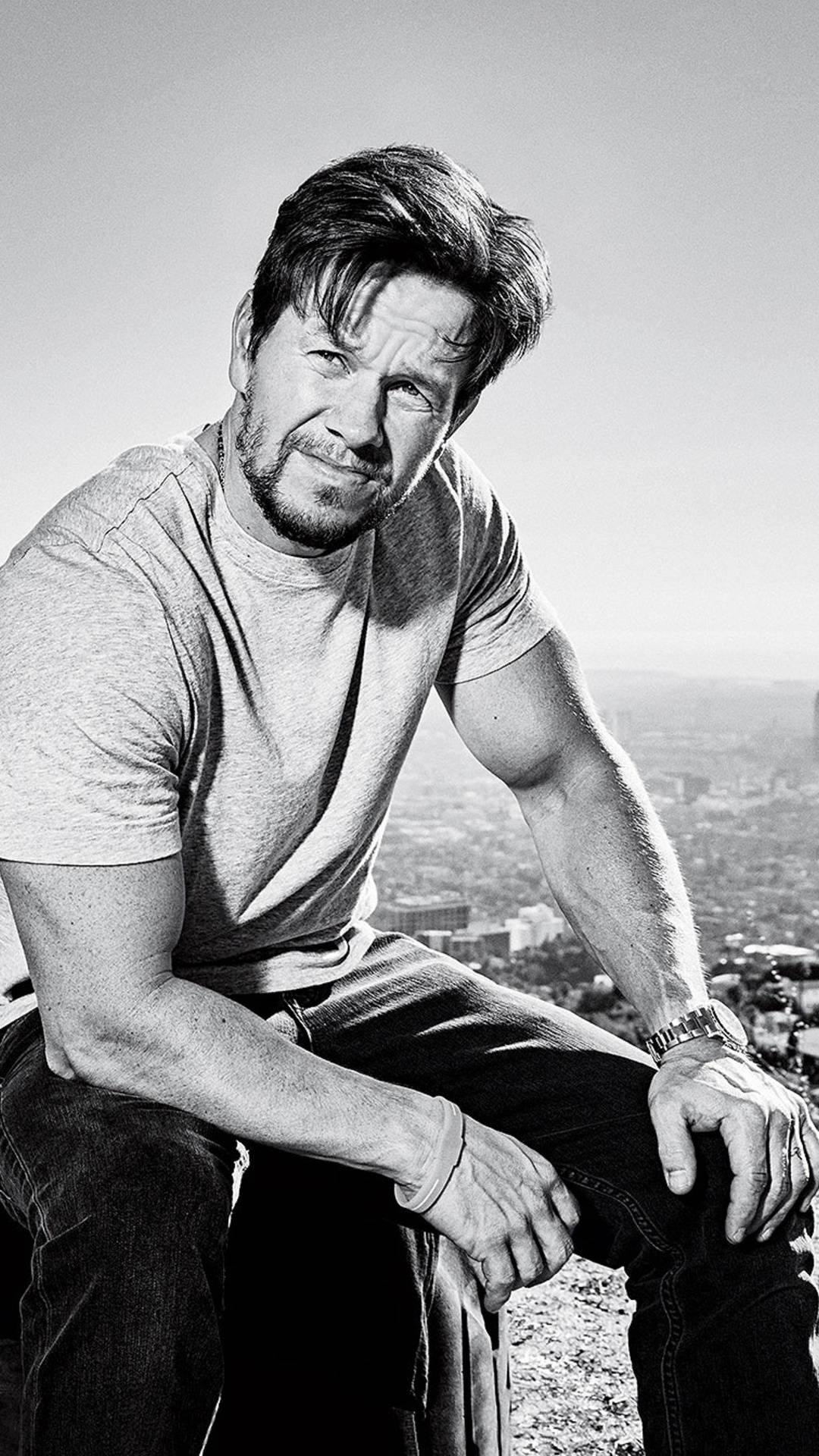 Mark Wahlberg 桌布 壁紙 — 一個留鬍子的男人坐在山頂上，俯瞰廣闊的城市景觀，是一幅戲劇性的黑白肖像。