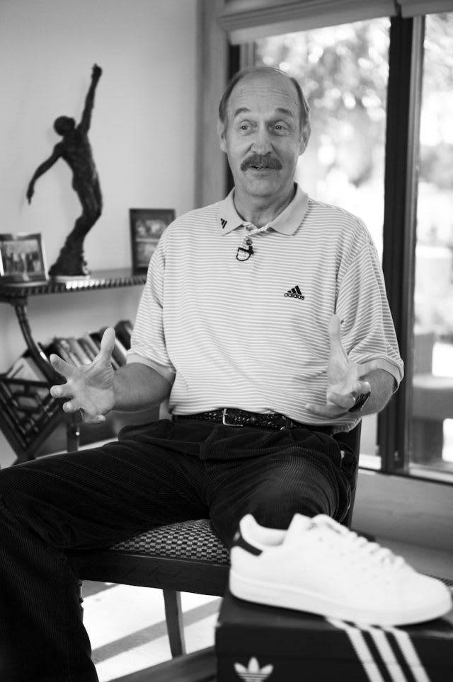 Shoe 桌布 壁紙 — 一個留鬍子的男人坐著做手勢，旁邊一個盒子上放著一雙白色 adidas 運動鞋，室內為單色調。