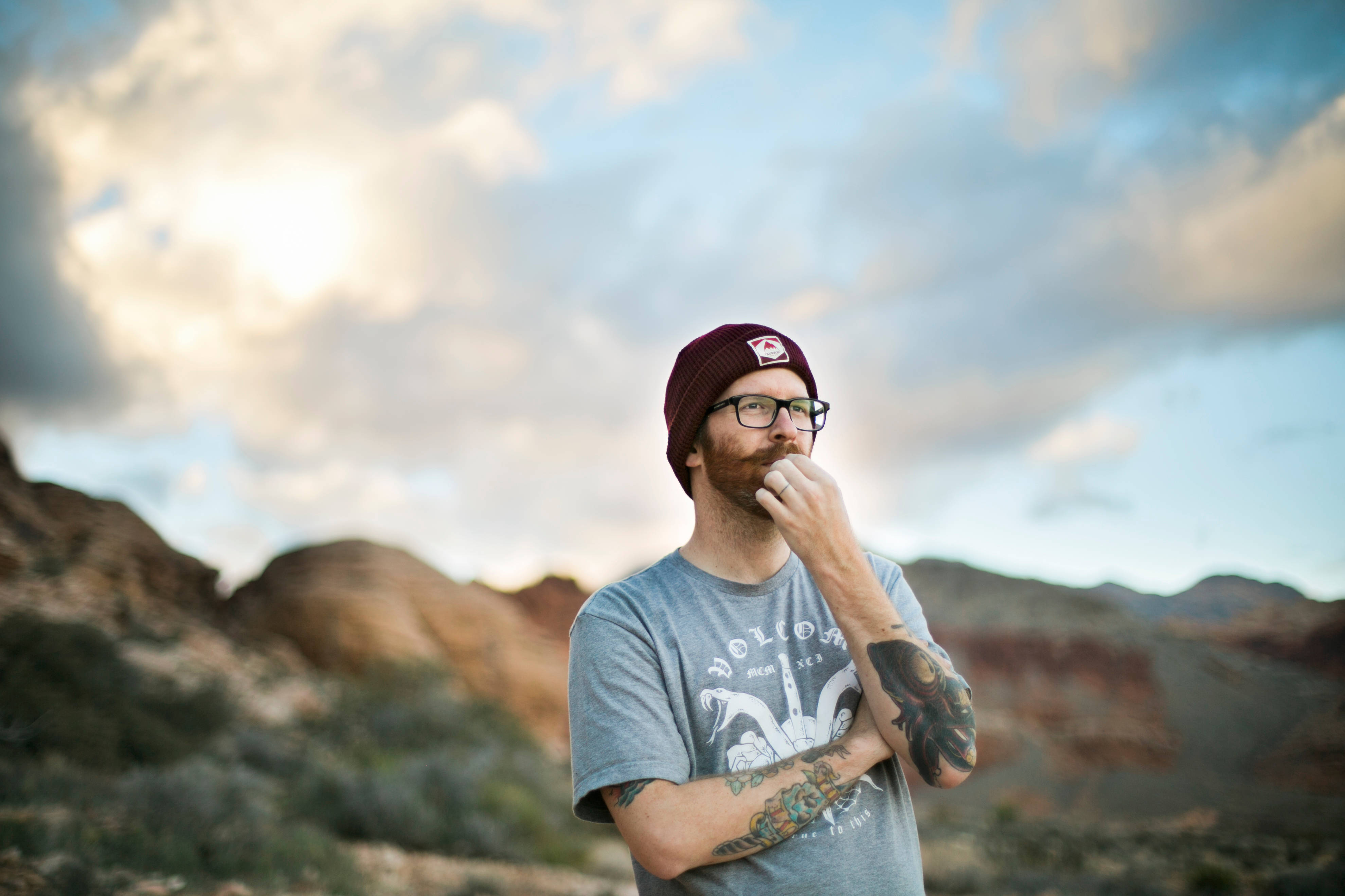 Hipster 桌布 壁紙 — 一個留鬍子的男人戴著栗色毛帽和眼鏡，在日落時分以戲劇性的沙漠峽谷為背景，若有所思地站著。