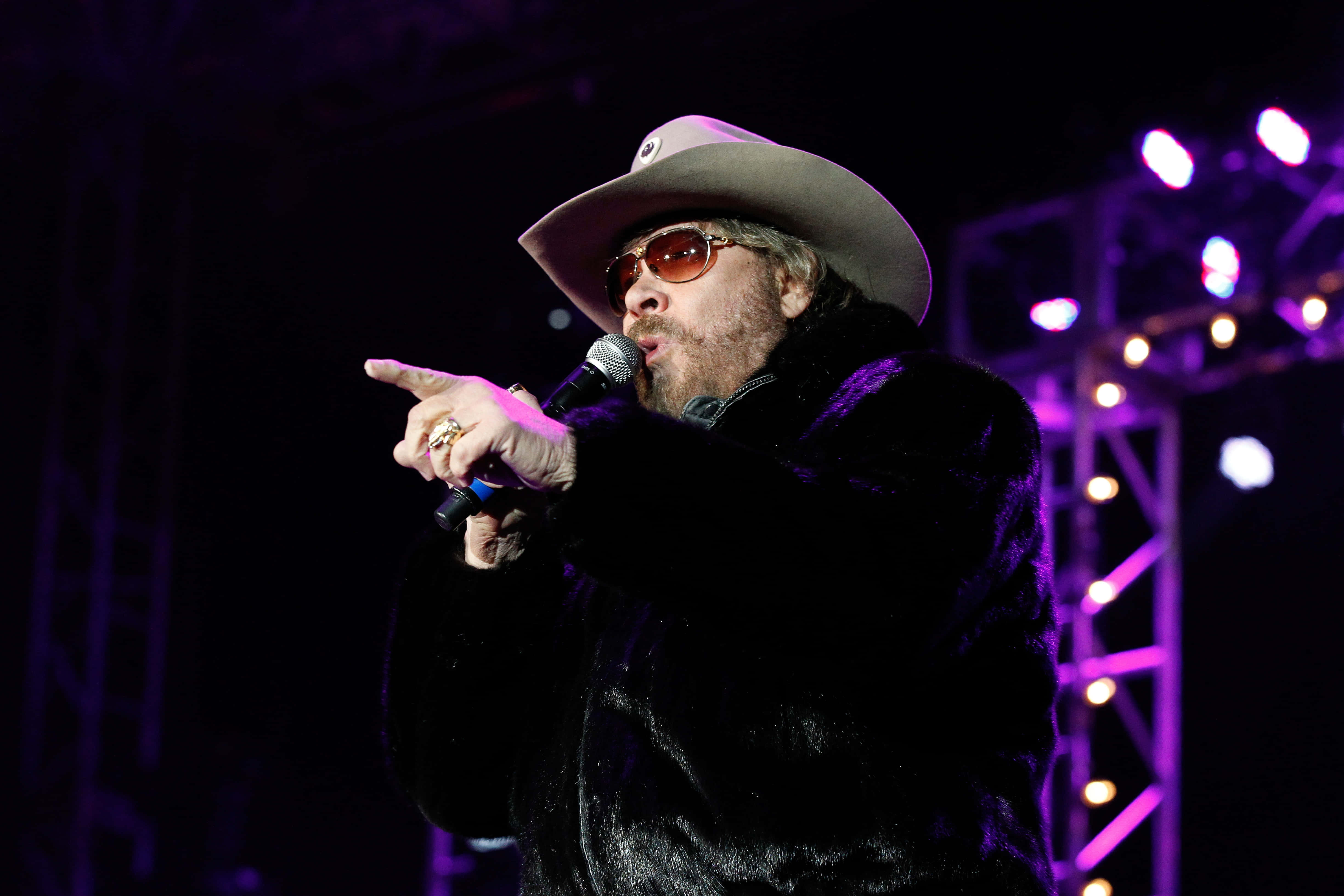 Hank Williams Jr 桌布 壁紙 — 一個留鬍子的男人戴著牛仔帽和太陽眼鏡，在紫色舞台燈光下對著麥克風唱歌。