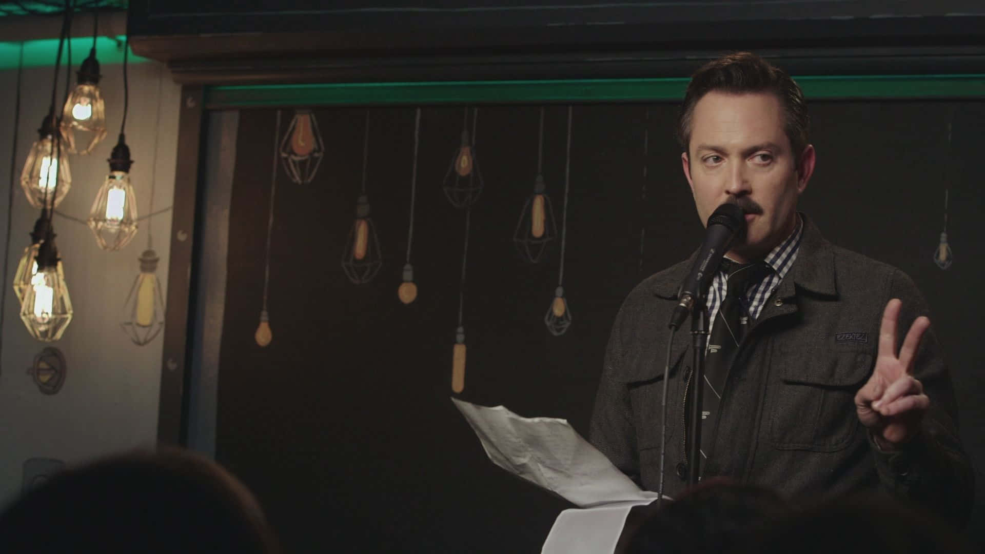 thomas lennon 桌布 壁紙 — 一個留鬍子的男人拿著文件，在懸掛的愛迪生燈泡下對著麥克風說話並比出和平手勢。