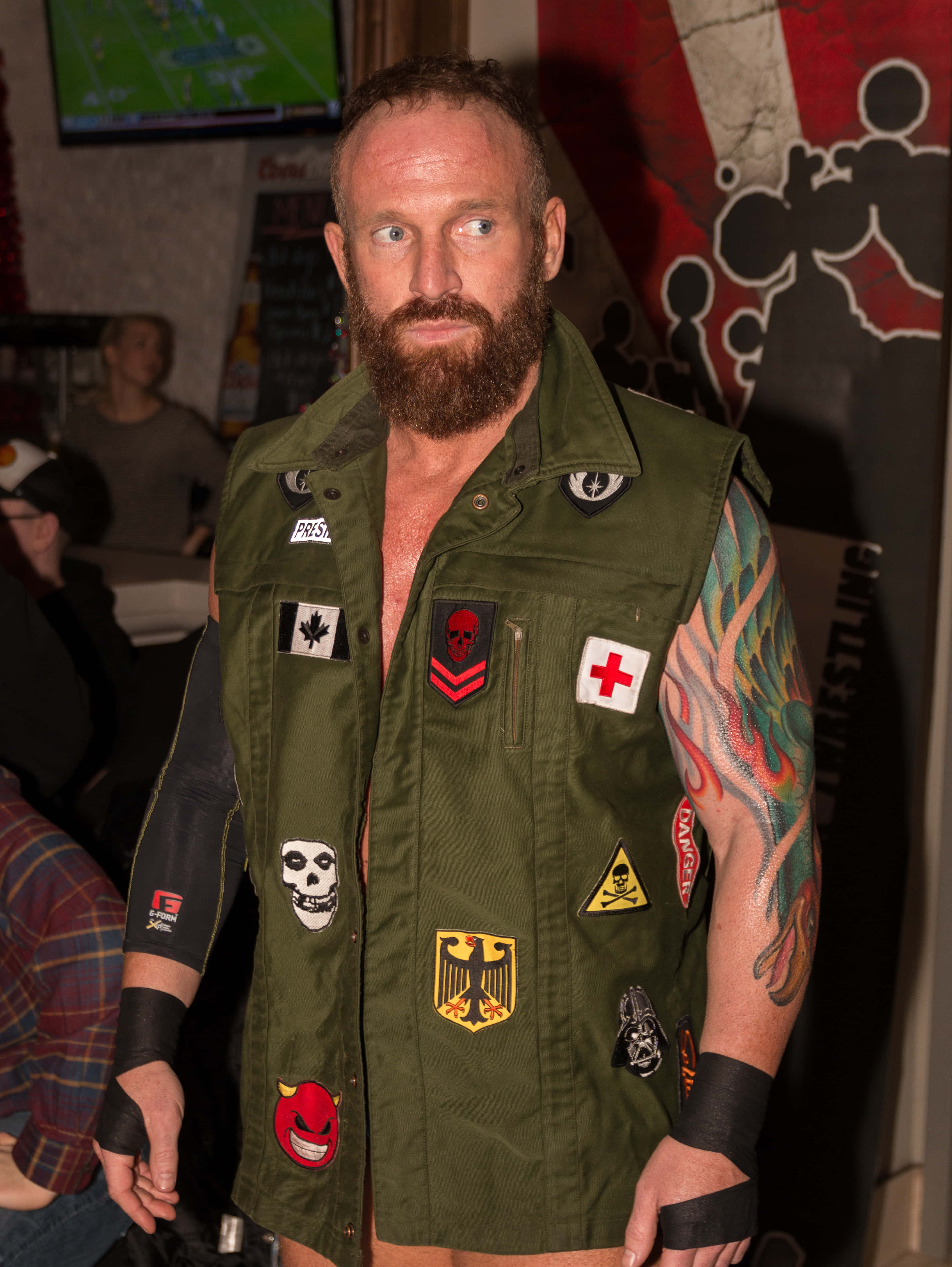 WWE 桌布 壁紙 — 一個留鬍子的男人穿著一件佈滿補丁和紋身的橄欖色背心，在昏暗的房間裡凝視著鏡頭。